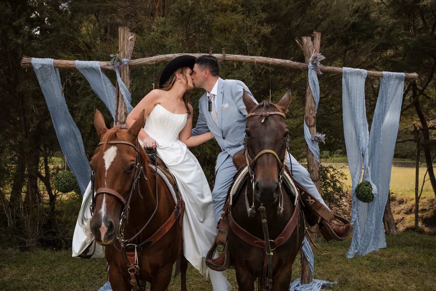 Bride and groom kissing on horseback under a rustic wooden arch adorned with blue fabric at an outdoor wedding.
