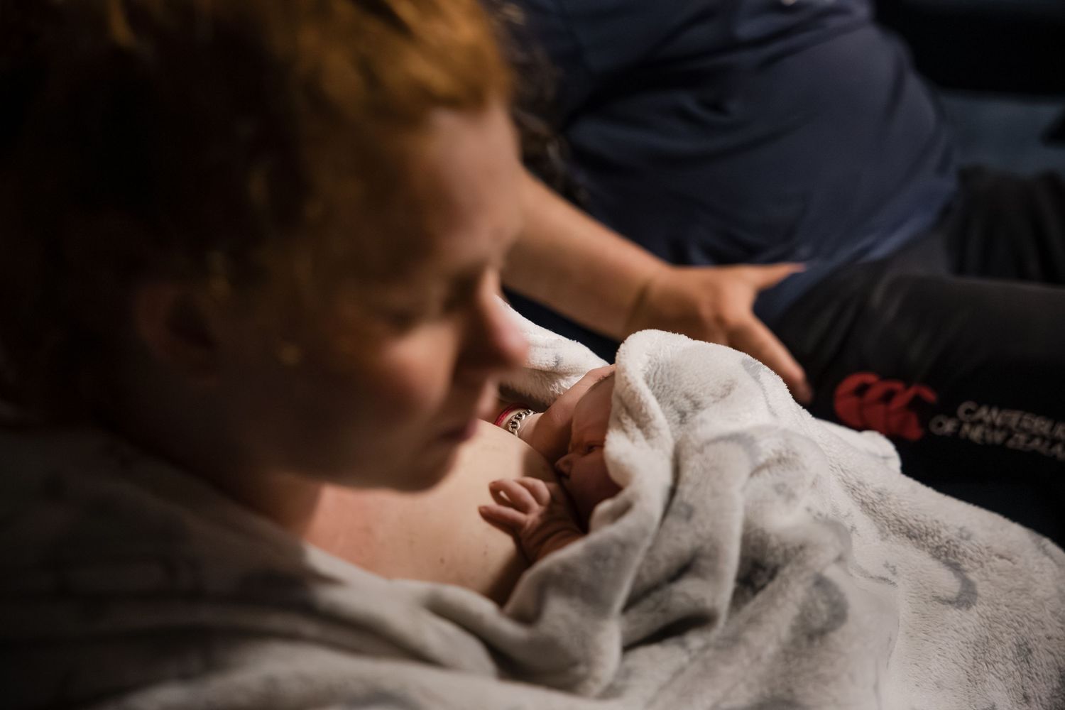 A parent and newborn share a tender moment together under a soft blanket in dim lighting.