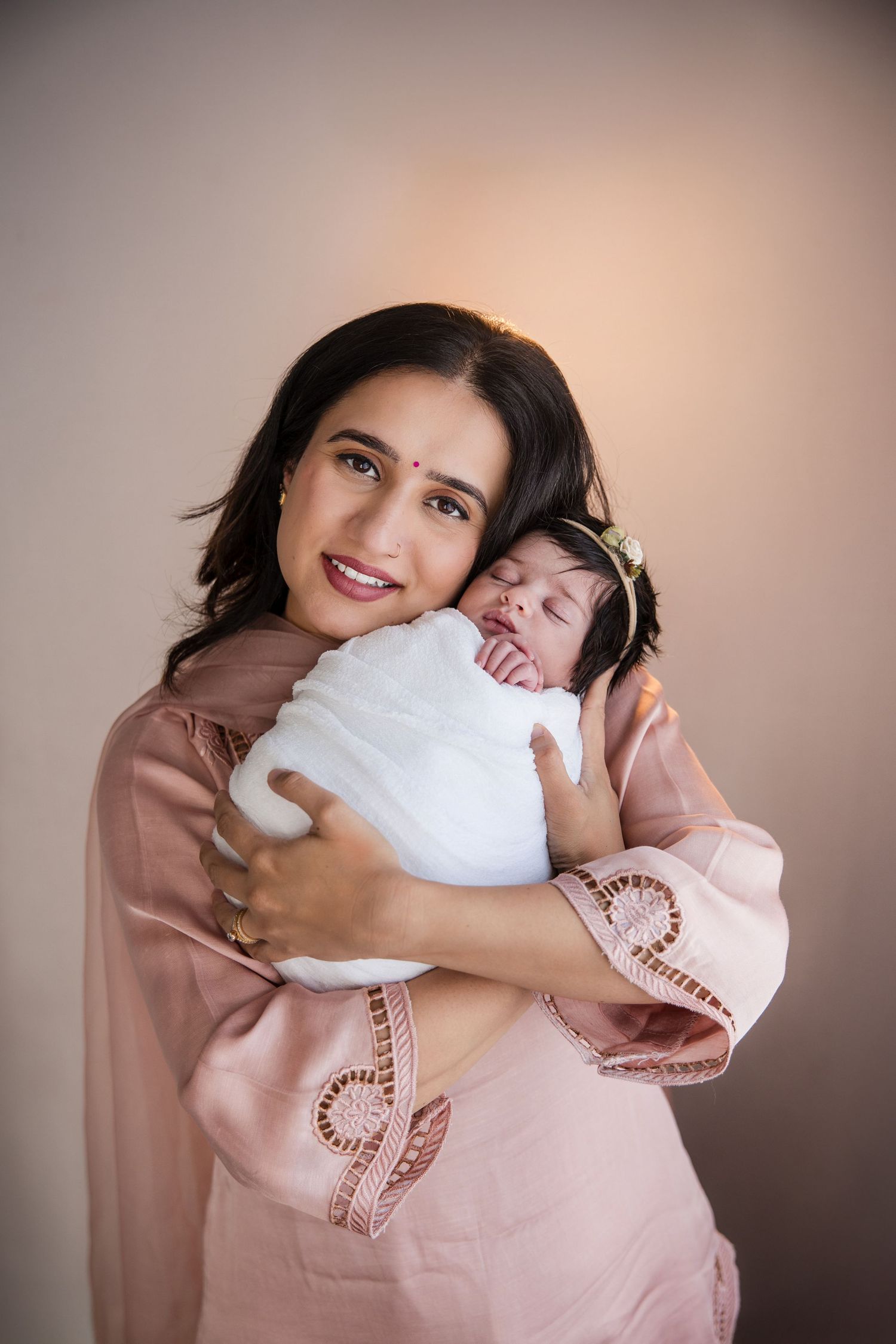 A smiling mother in a pink embroidered dress tenderly holds her sleeping newborn baby wrapped in white.