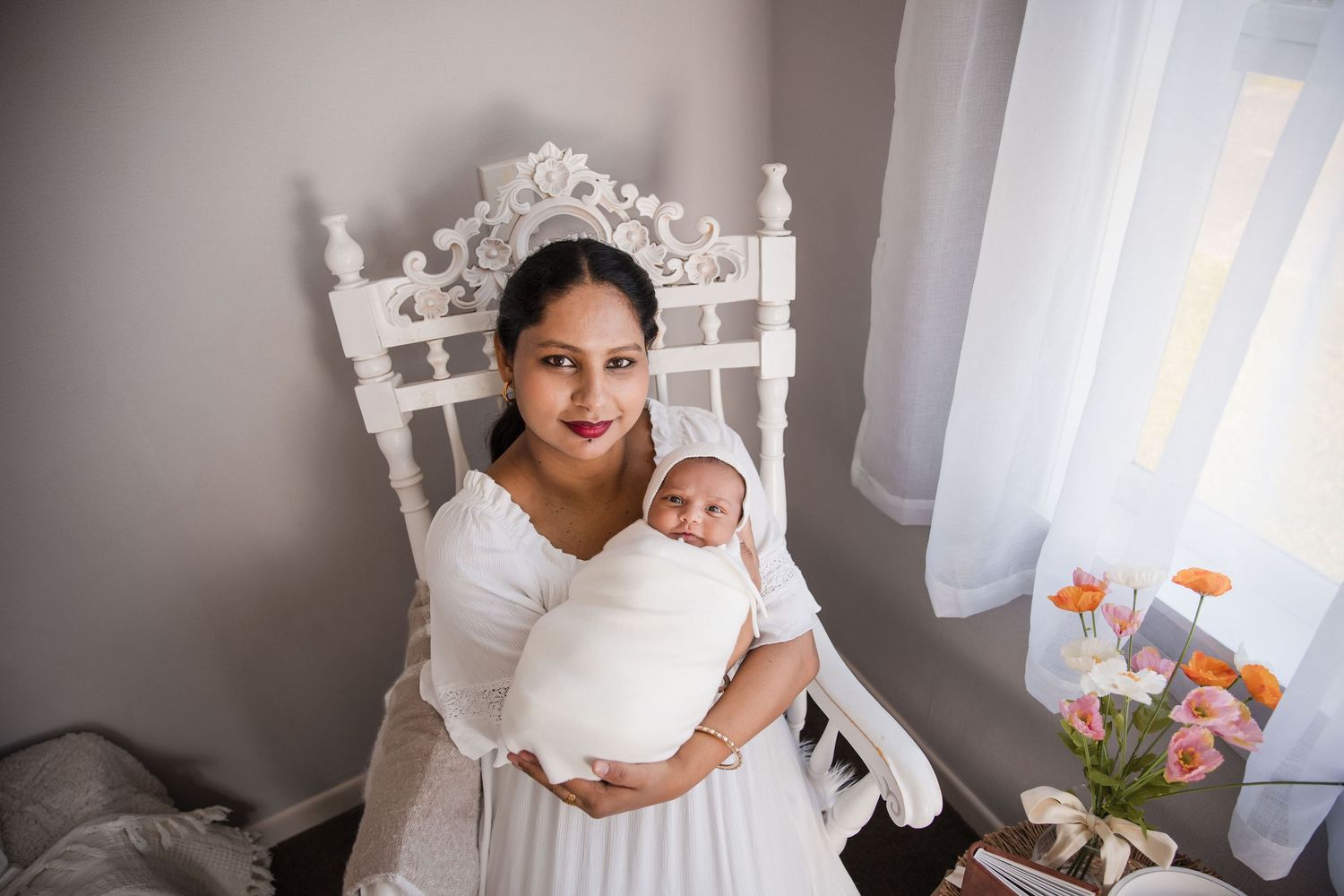 A mother in a white dress holds her swaddled newborn baby while seated in an ornate white chair near a window.