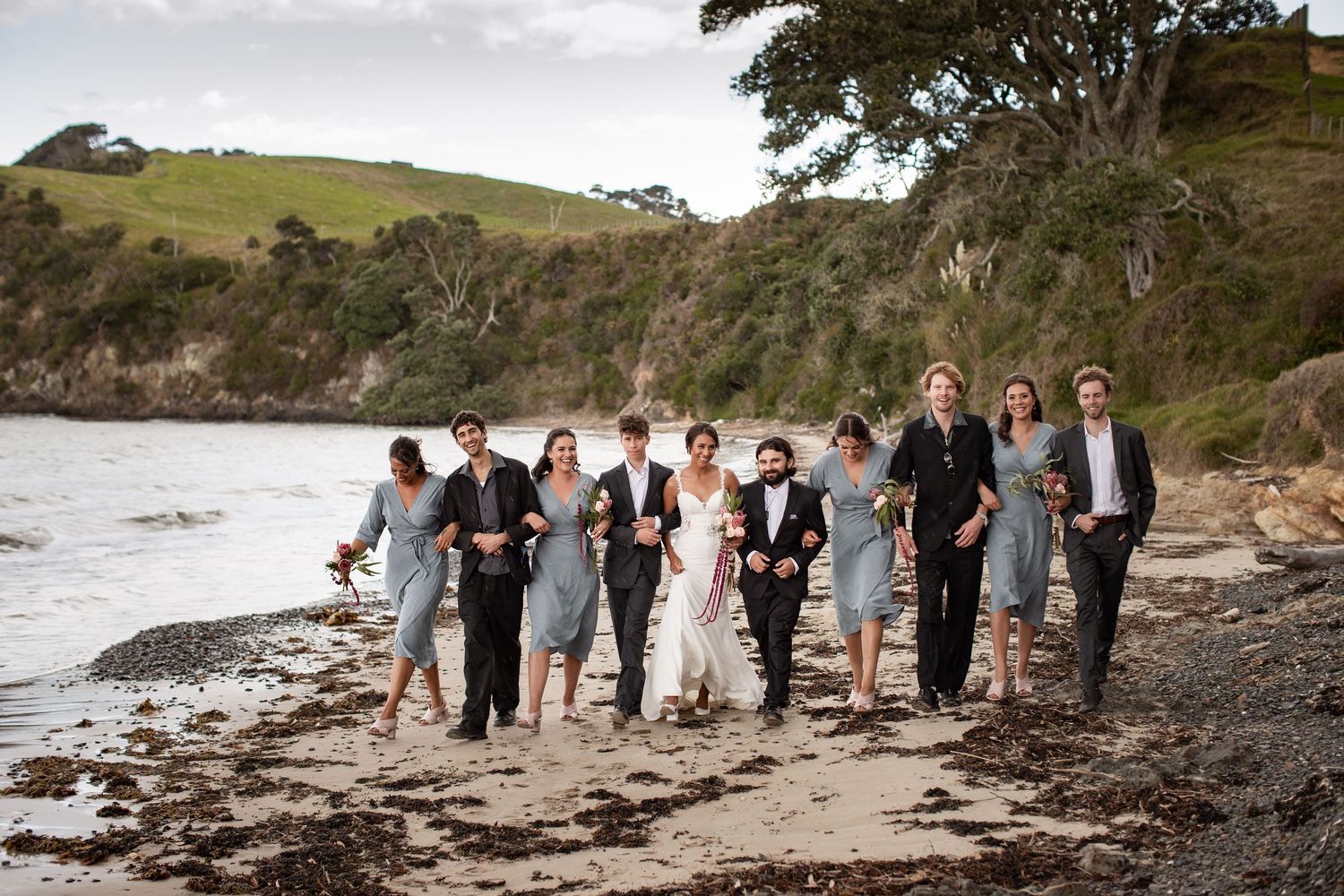 A group walking and laughing together along a scenic rocky beach with green hills in the background.