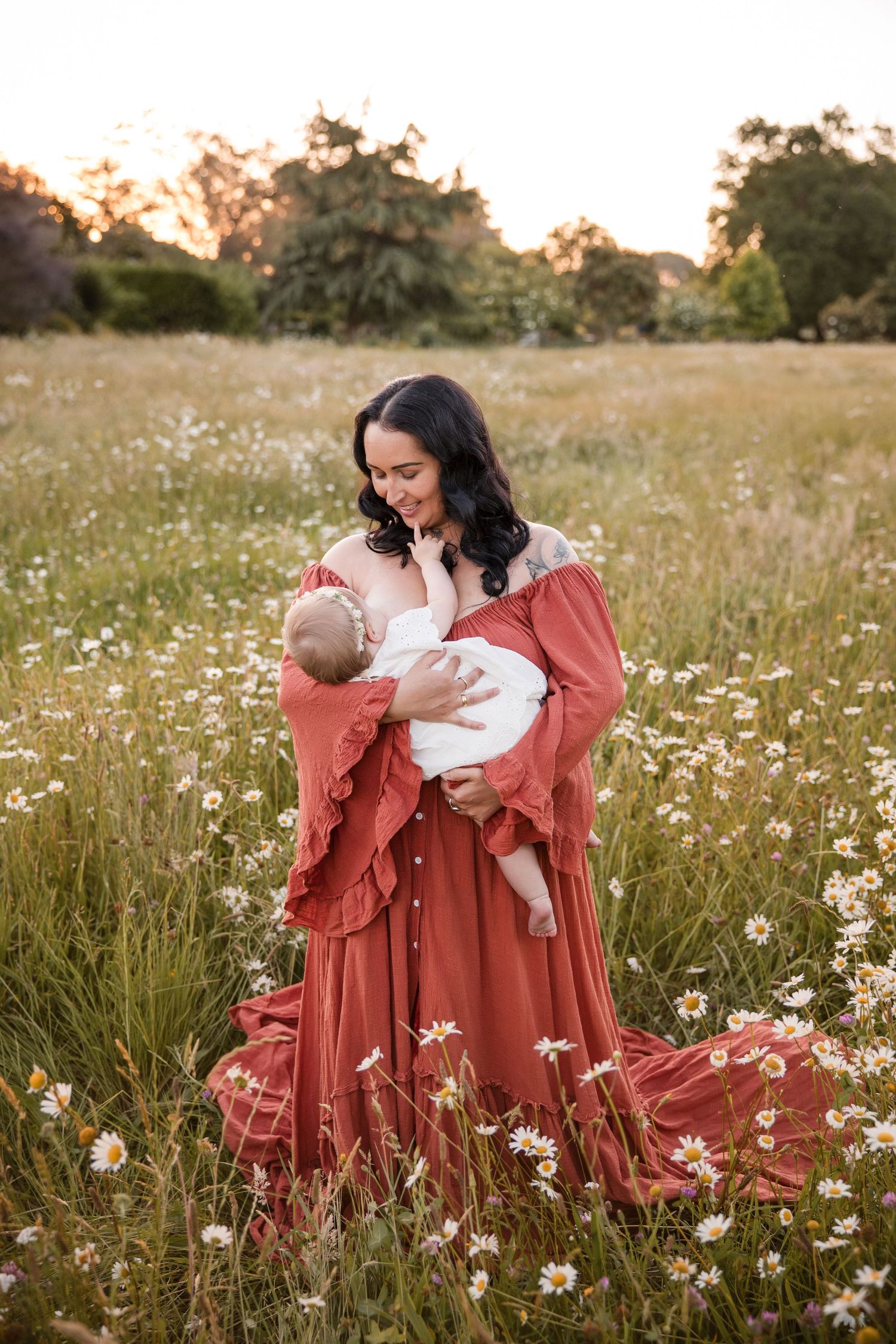 A smiling mother in a rust red dress holds her newborn baby in a sunlit wildflower meadow at golden hour.