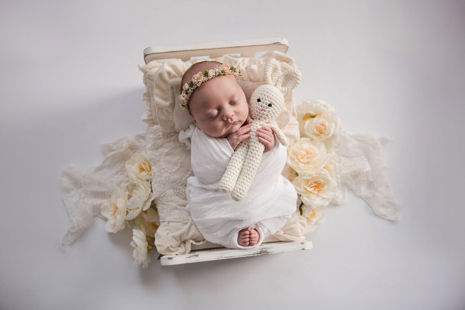 Newborn baby wrapped in white swaddle sleeping in a wooden crib with cream roses and floral headband holding a knit toy.
