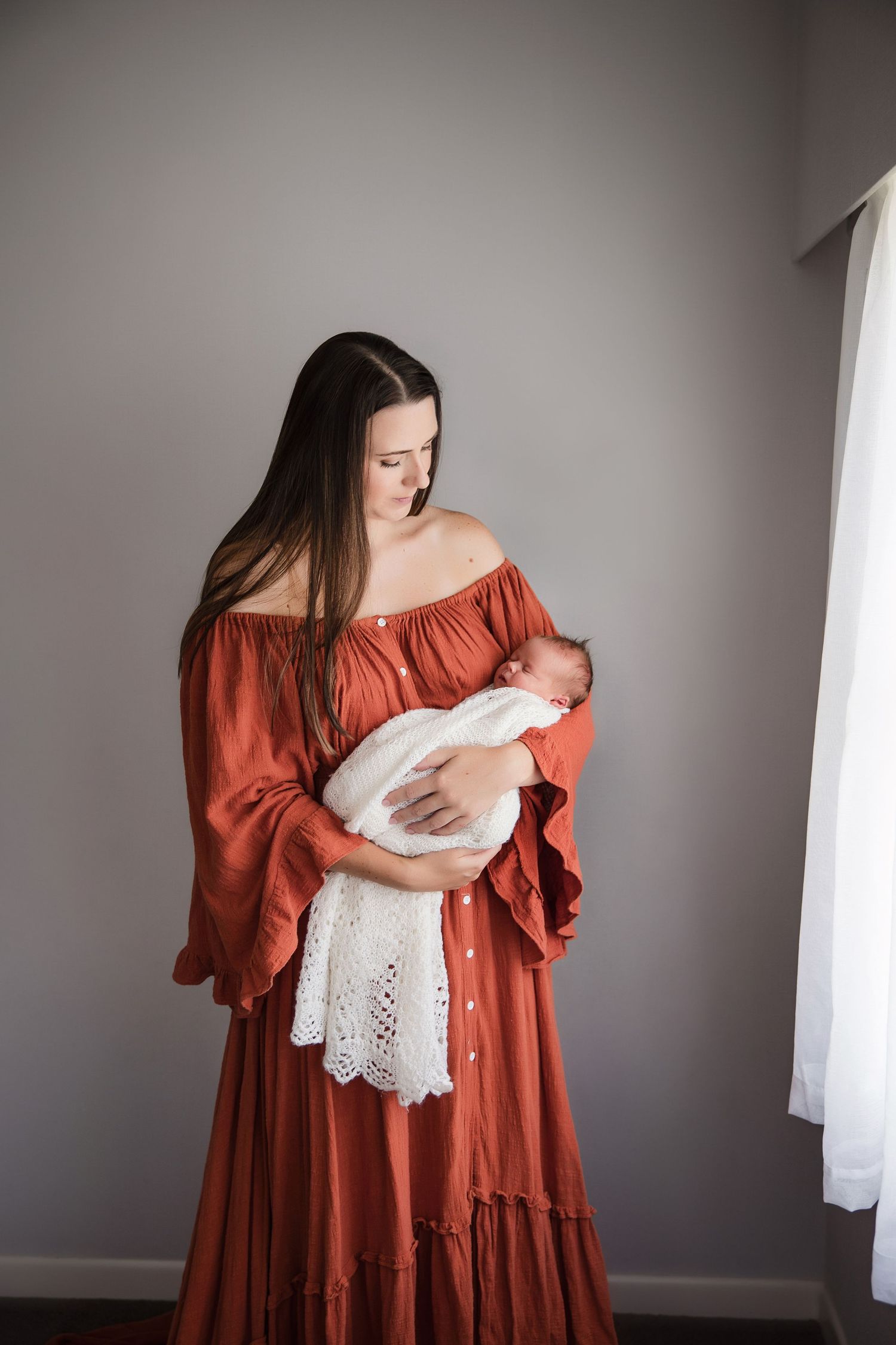 A young mother in a rust-colored off-shoulder dress tenderly holds her sleeping newborn wrapped in white lace.