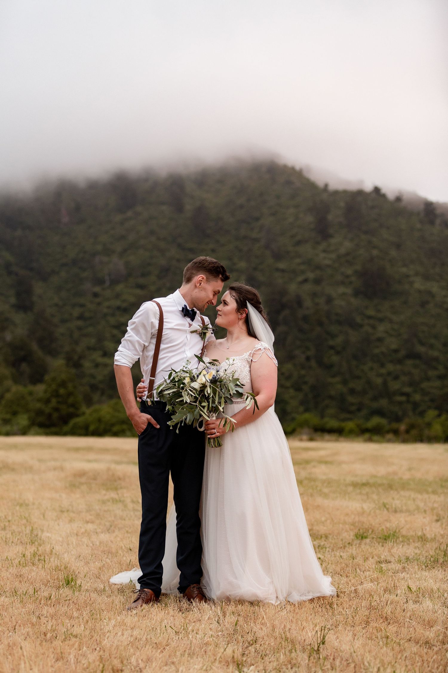 A couple shares a romantic moment in their wedding attire against a misty mountain backdrop in a golden field.