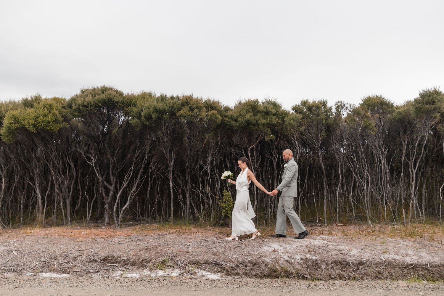 A couple hold hands while walking in nature with a backdrop of dark trees on an overcast day.