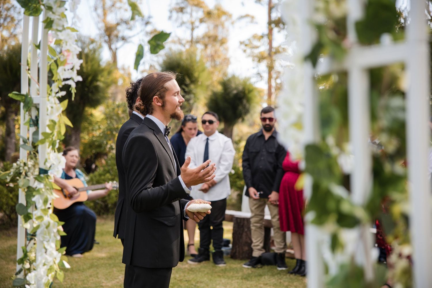 A speaker delivers remarks during an outdoor garden wedding ceremony surrounded by greenery and natural decor.