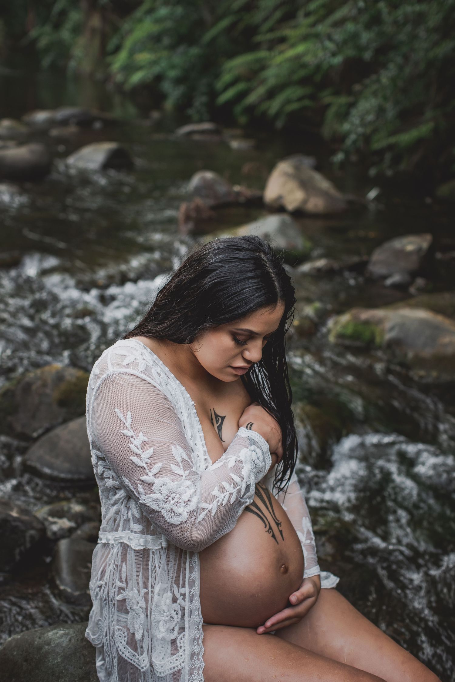 Serene maternity portrait by a rocky stream showing white lace dress against natural backdrop.