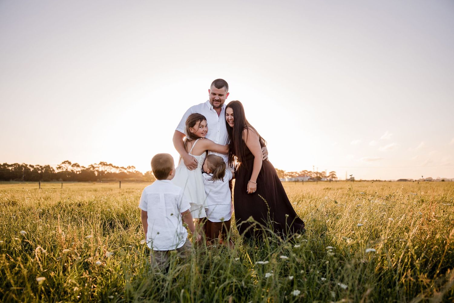 Family of five posing in golden field at sunset, parents with three young children in warm natural light.