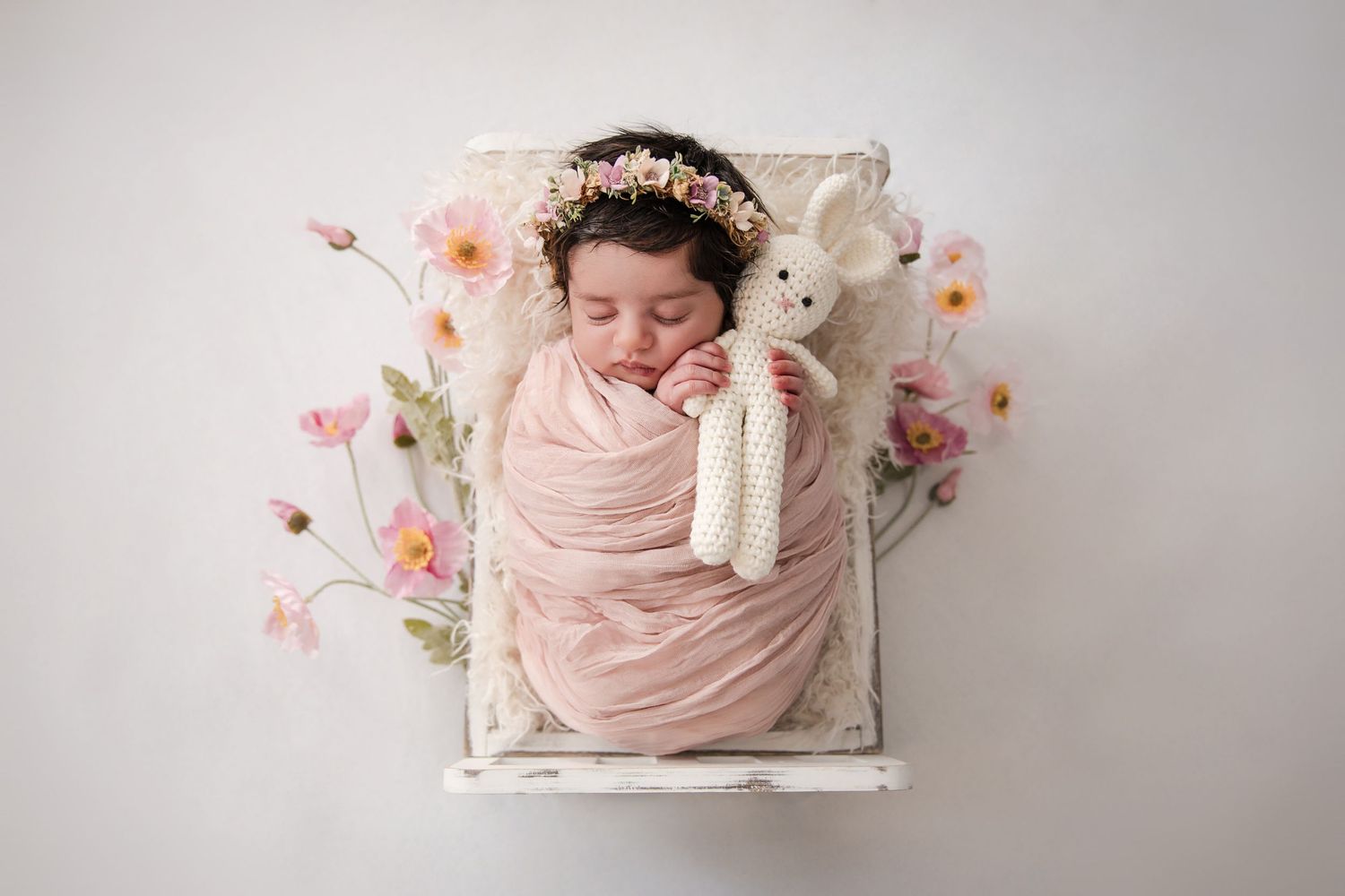 Newborn baby swaddled in pink wrap sleeping in white mini bed with floral crown and knit bunny toy.