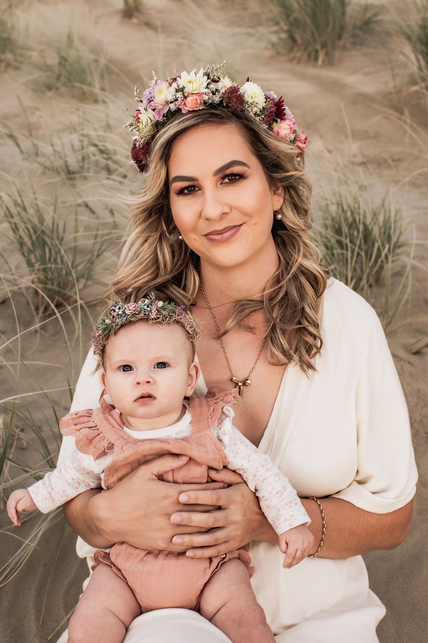 Mother and baby wearing floral crowns pose together on sandy beach dunes at sunset.