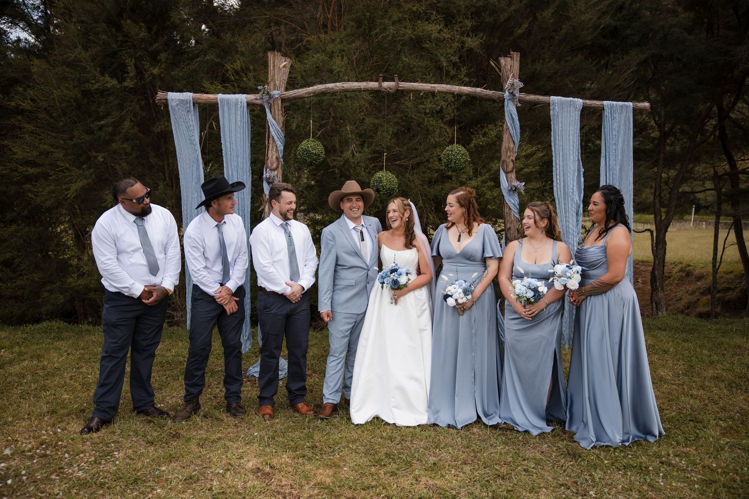Wedding party poses outdoors under rustic wooden arch with blue fabric, bridesmaids in dusty blue gowns, groomsmen in gray suits.