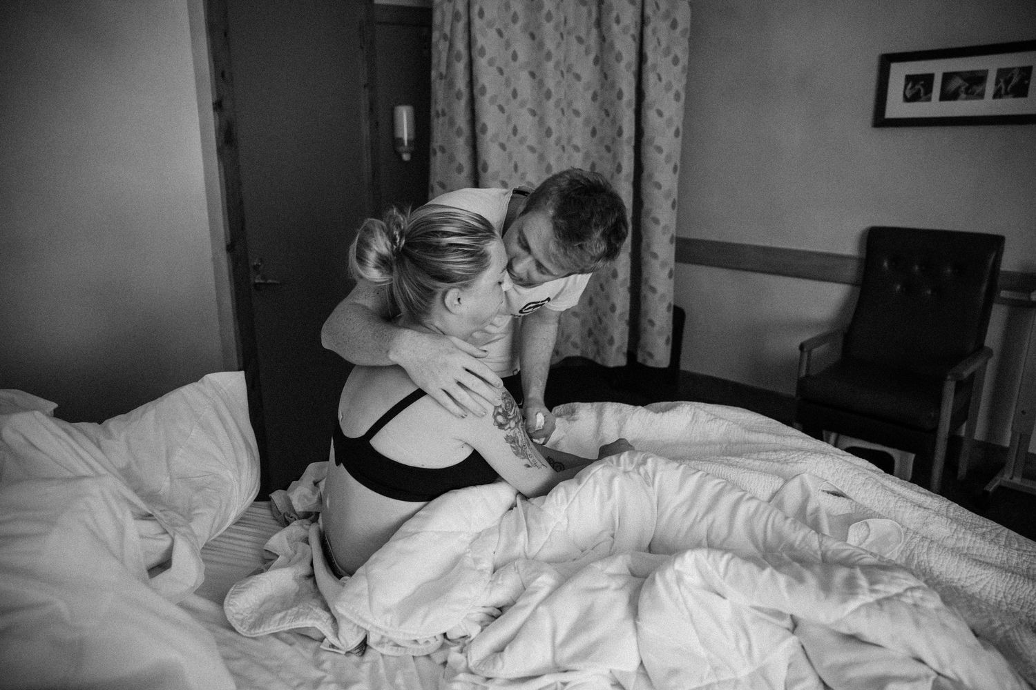 Black and white photo of a pregnant couple sharing an intimate embrace on a bed during labor.