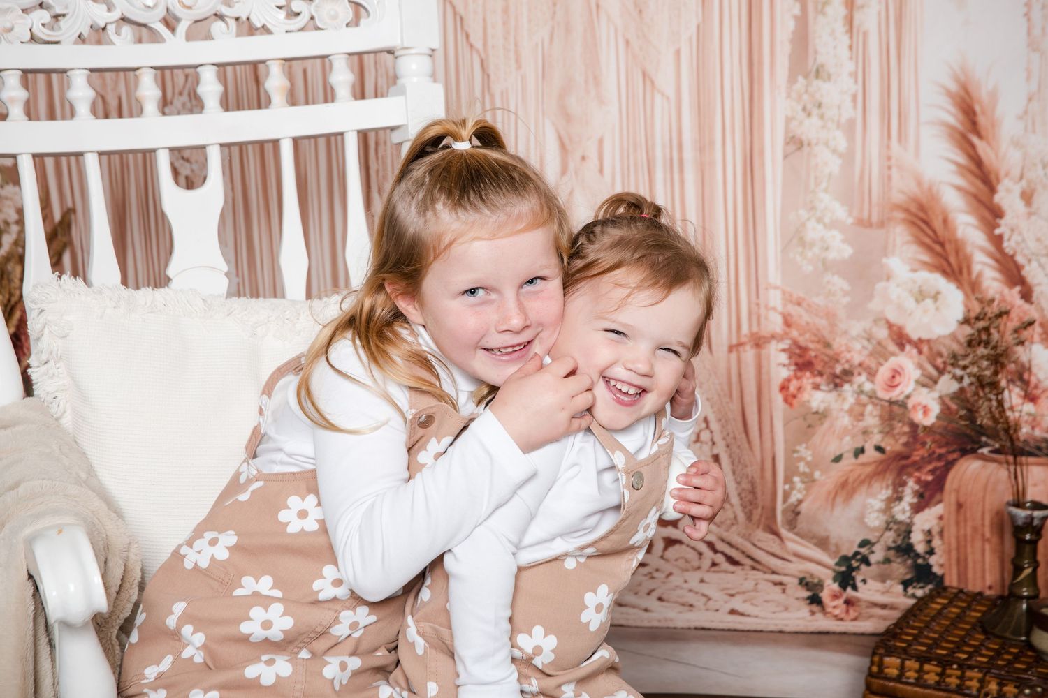Two children in white clothing share a joyful embrace while sitting on a floral-patterned chair with pink decor.