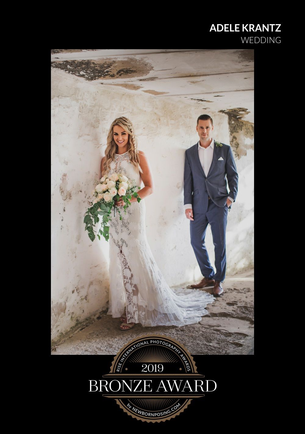 A bride in a lace gown and groom in a gray suit pose against a rustic white wall with a Bronze Award emblem below.