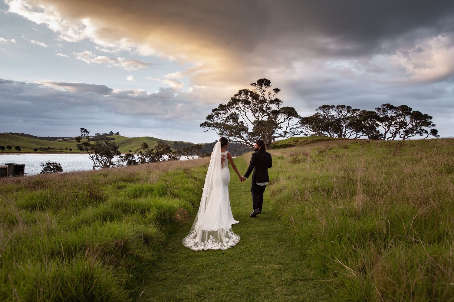 Couple stands together on grassy hillside overlooking water at sunset with dramatic cloudy sky.