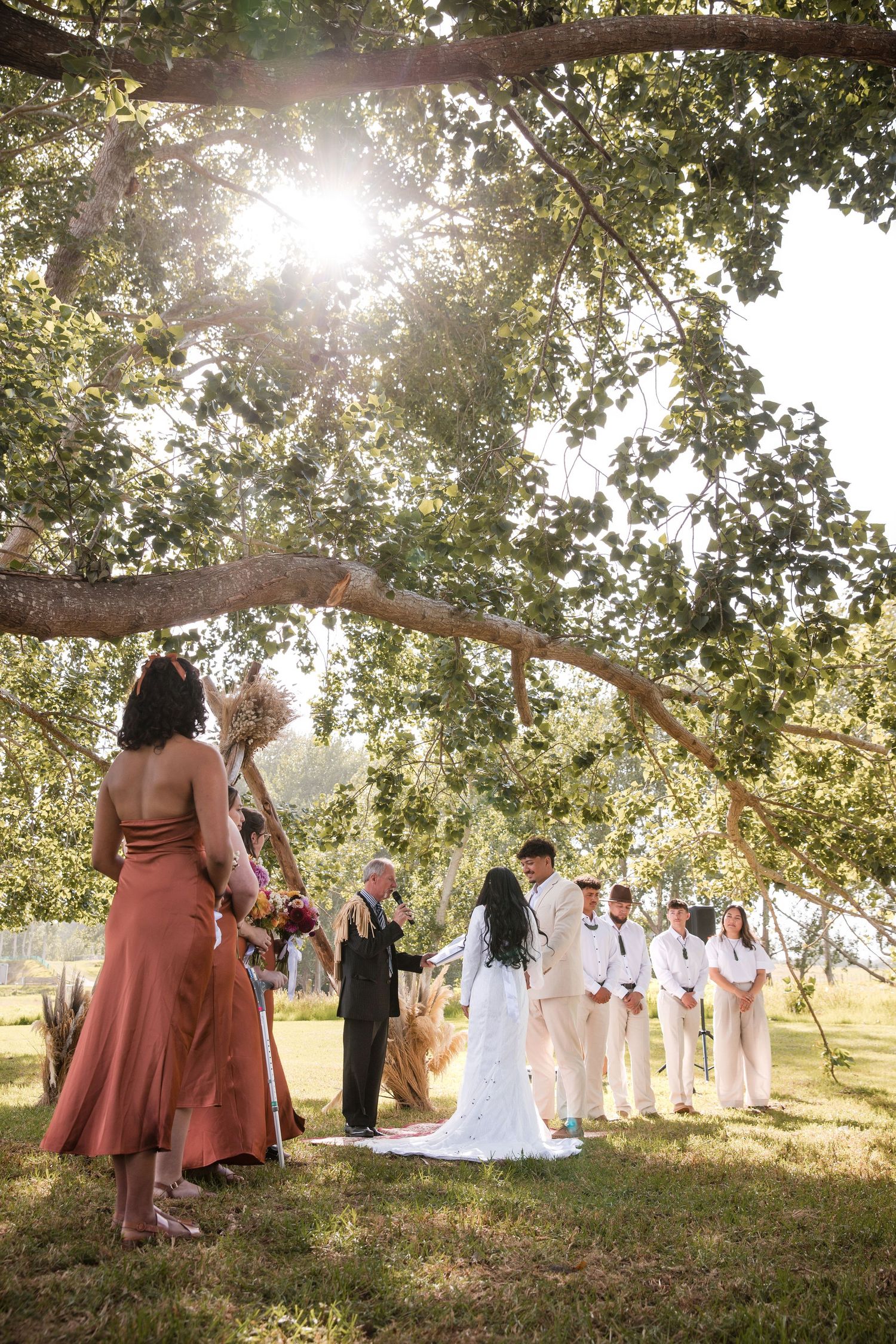 An outdoor wedding ceremony under a large tree with sunlight streaming through the leaves and bridal party in white attire.
