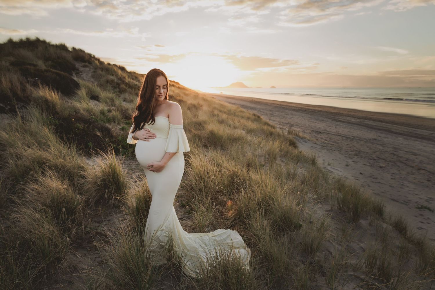 Pregnant woman in fitted dress poses on beach dunes during golden sunset. Cambridge Waikato NZ Photographer. White Locket Photography