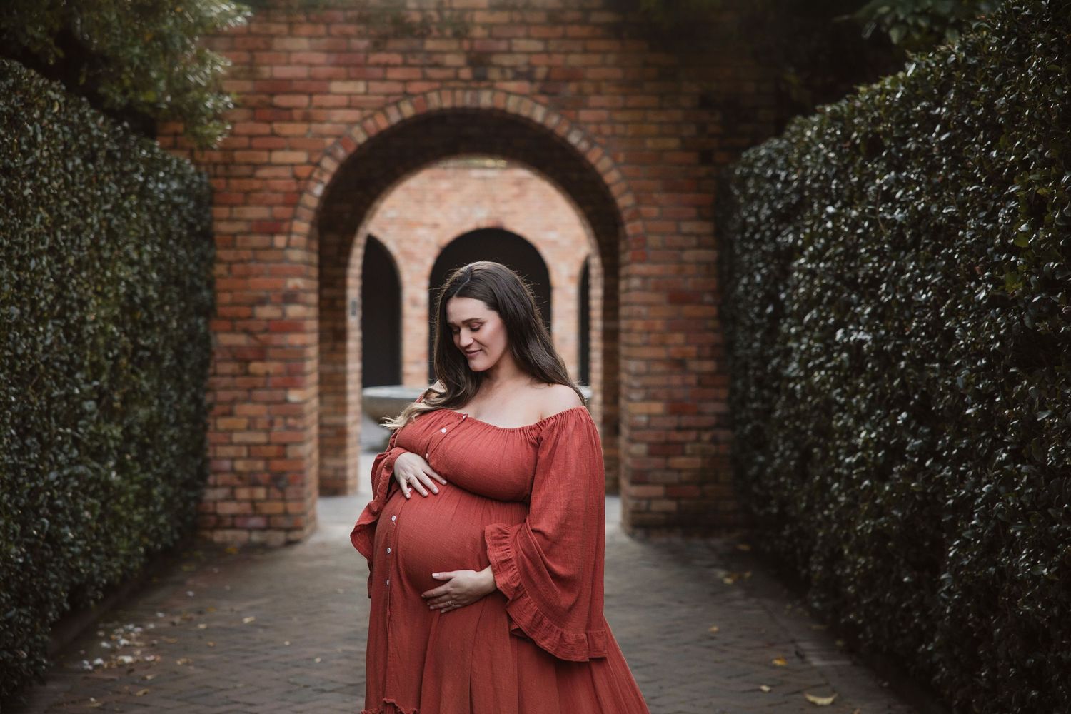 Pregnant woman in rust-colored off-shoulder gown cradling belly, standing in brick archway surrounded by lush hedges.