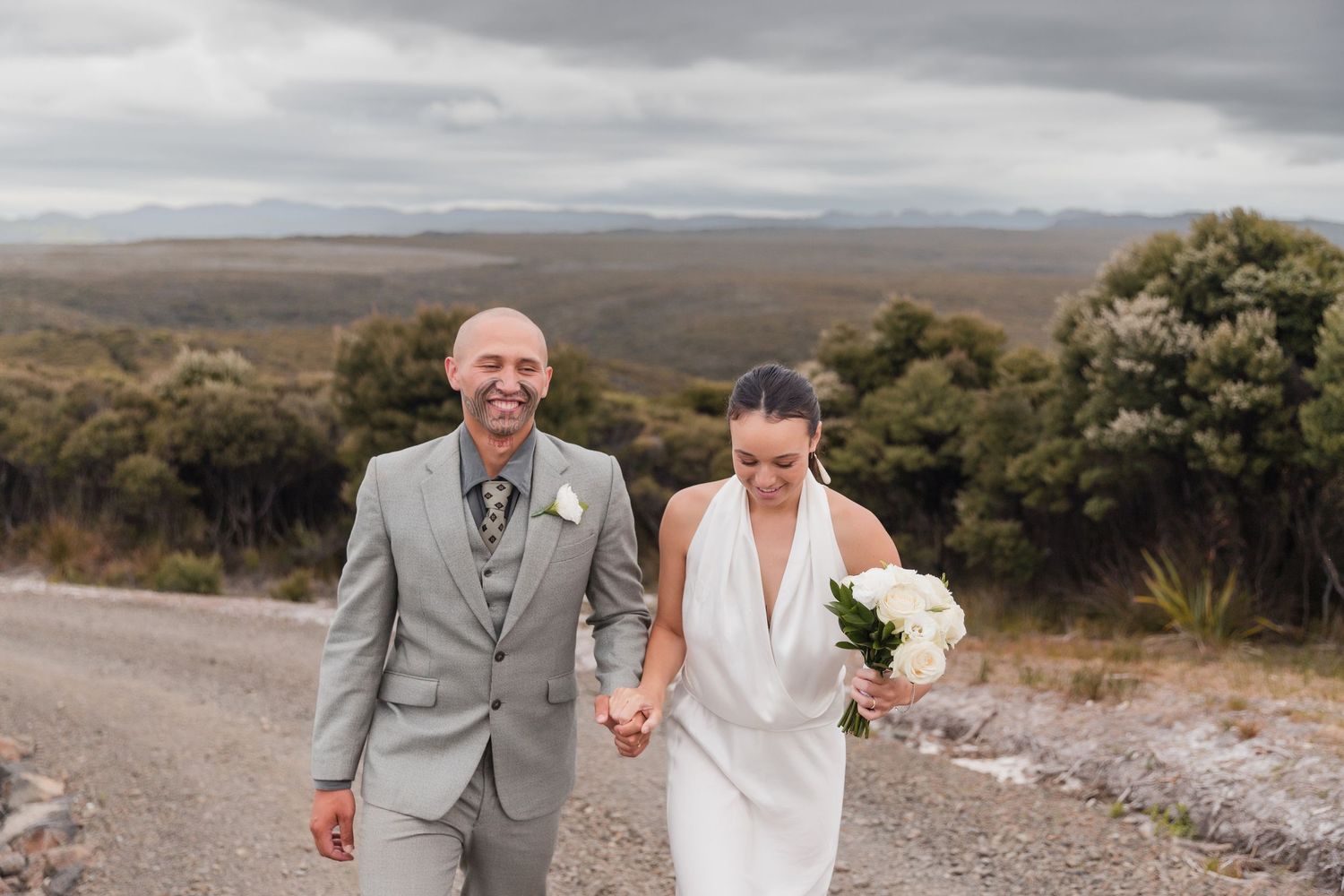 A couple in wedding attire walks on a mountain road with a scenic landscape view behind them.