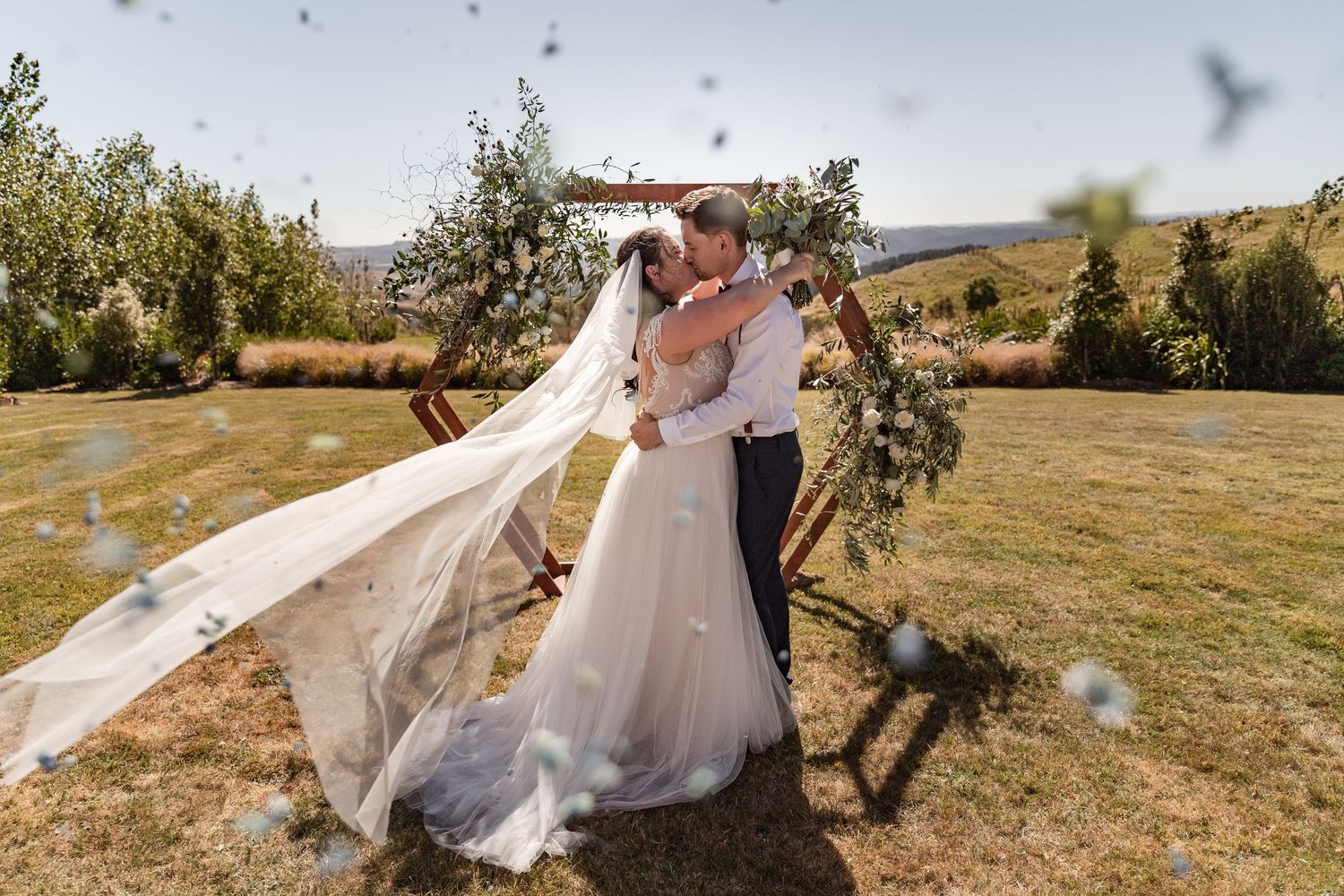 A couple embraces during an outdoor wedding ceremony as the bride's veil dramatically flows in the wind.