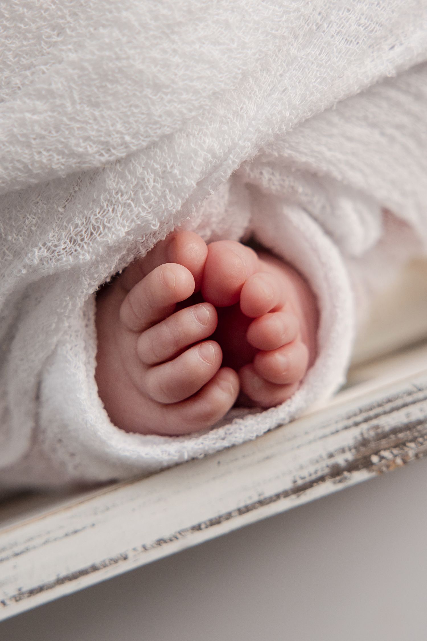 Close-up of tiny newborn baby feet peeking out from a soft white blanket on a rustic white wooden surface.