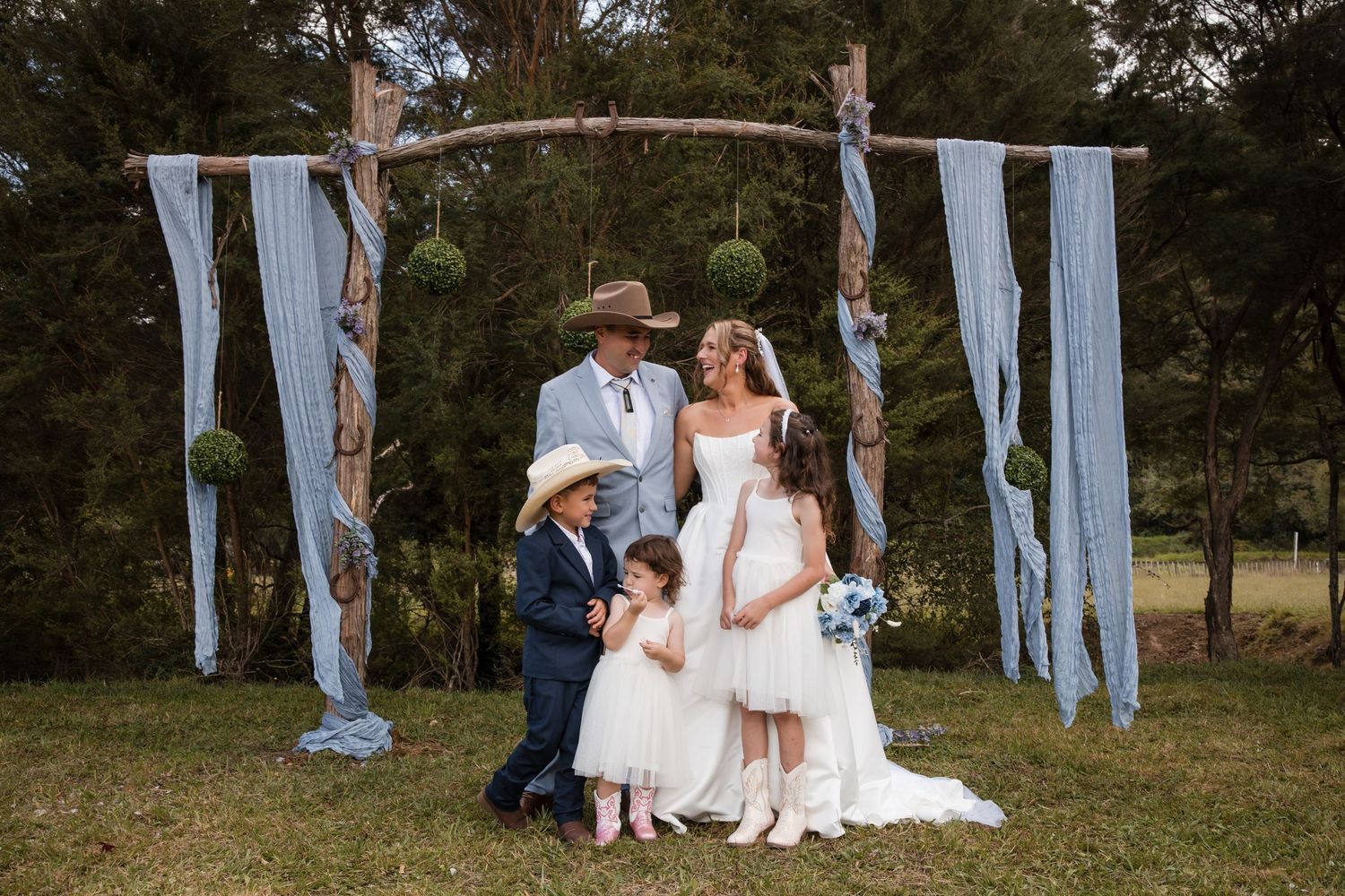 Bride and groom with children pose under rustic wooden arch with blue draping and green floral balls at outdoor wedding.