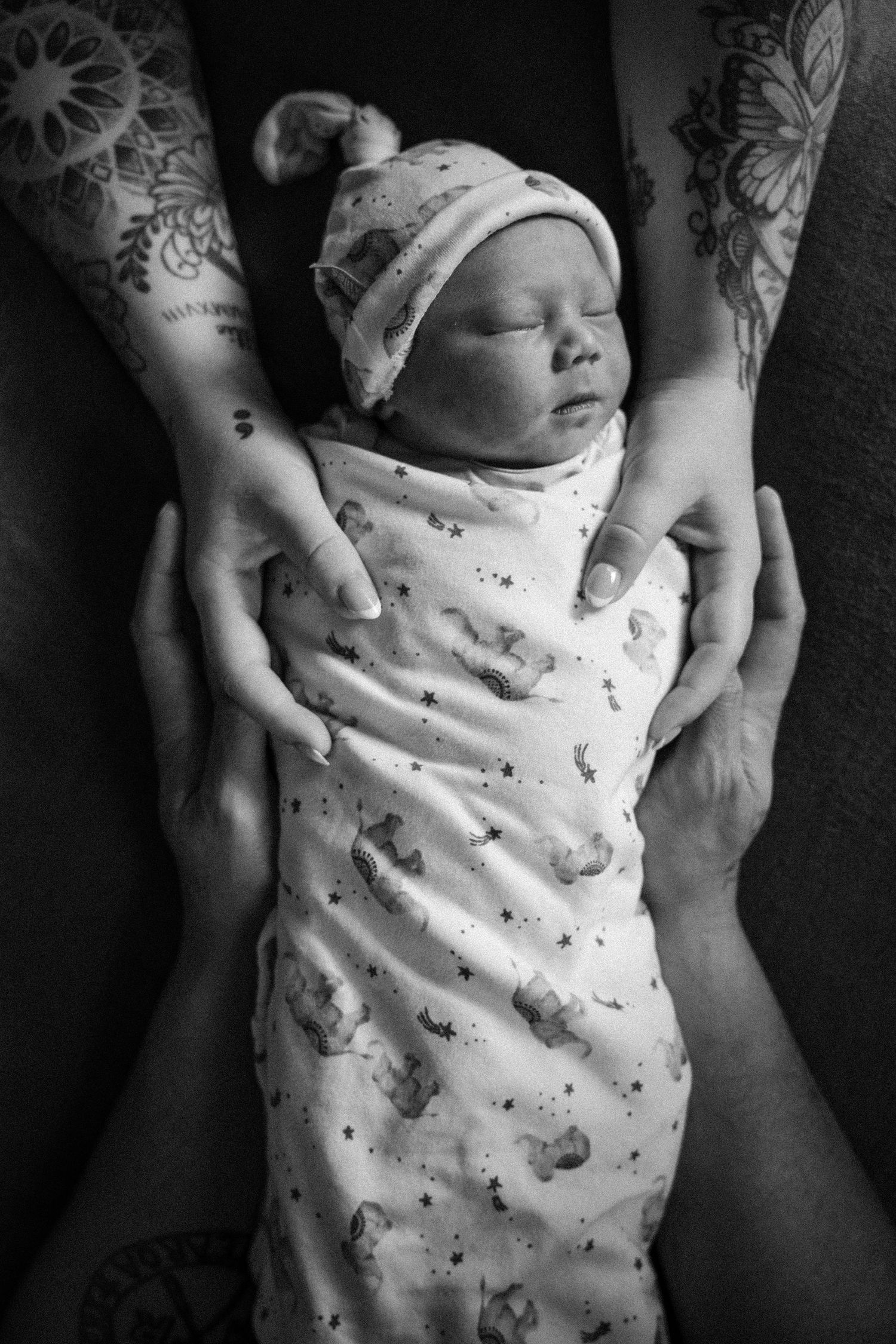 Black and white photo of a sleeping newborn baby swaddled in a patterned blanket, cradled by tattooed adult hands.