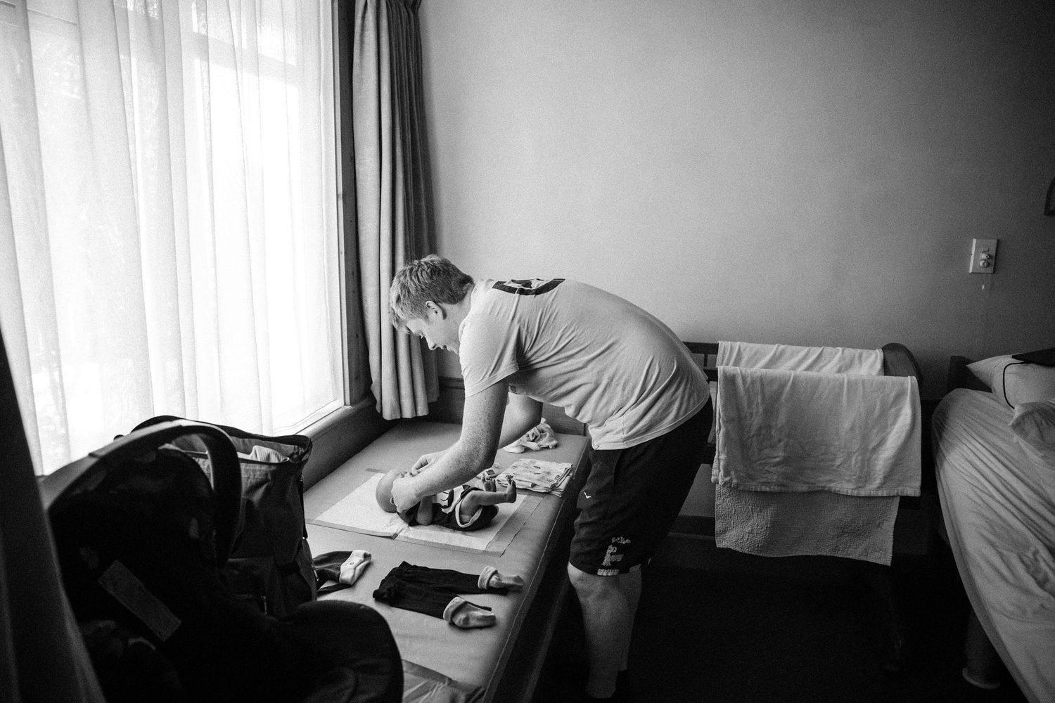 Black and white photo of a person organizing items on a table in a hotel room near a window.