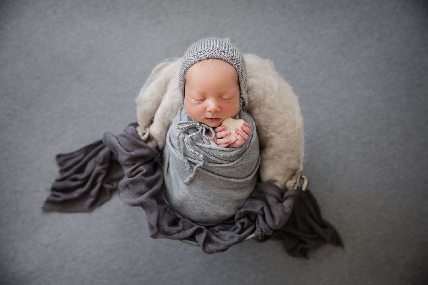 Sleeping newborn baby wrapped in gray swaddle with knit hat, nestled in wooden bowl on gray backdrop.