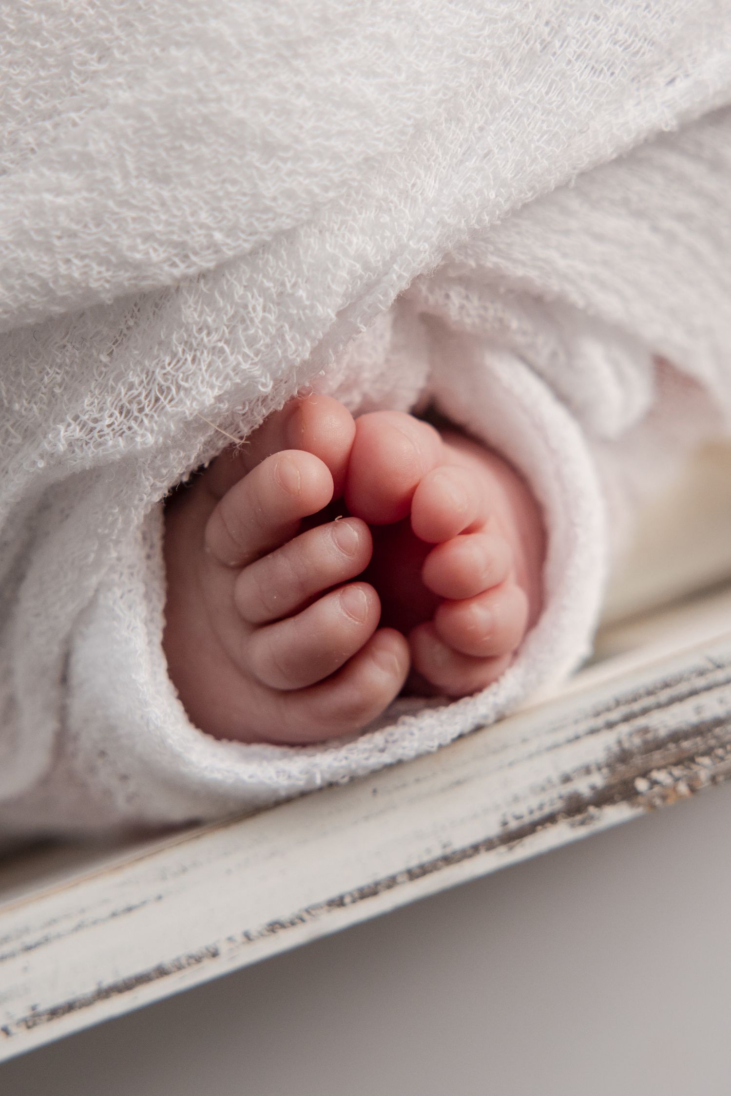 Tiny newborn baby feet peeking out from soft white blanket, resting on rustic white wooden surface.