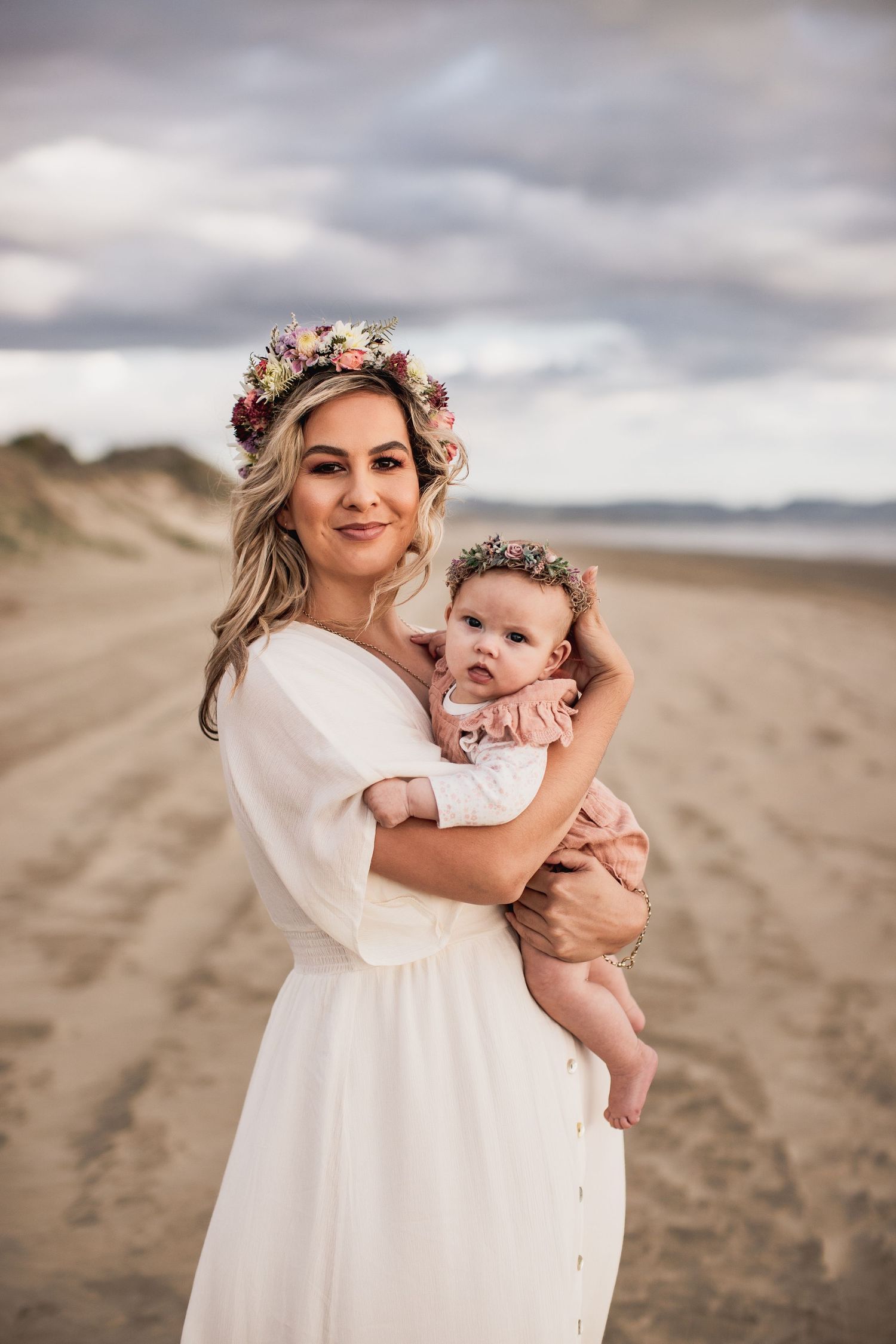 Beach portrait of mother and baby wearing matching floral crowns and white dresses.