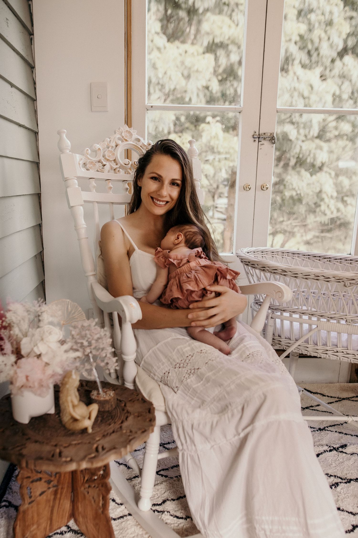 Woman in white dress sits in vintage rocking chair on porch holding baby in pink outfit. Cambridge Waikato NZ Photographer. White Locket