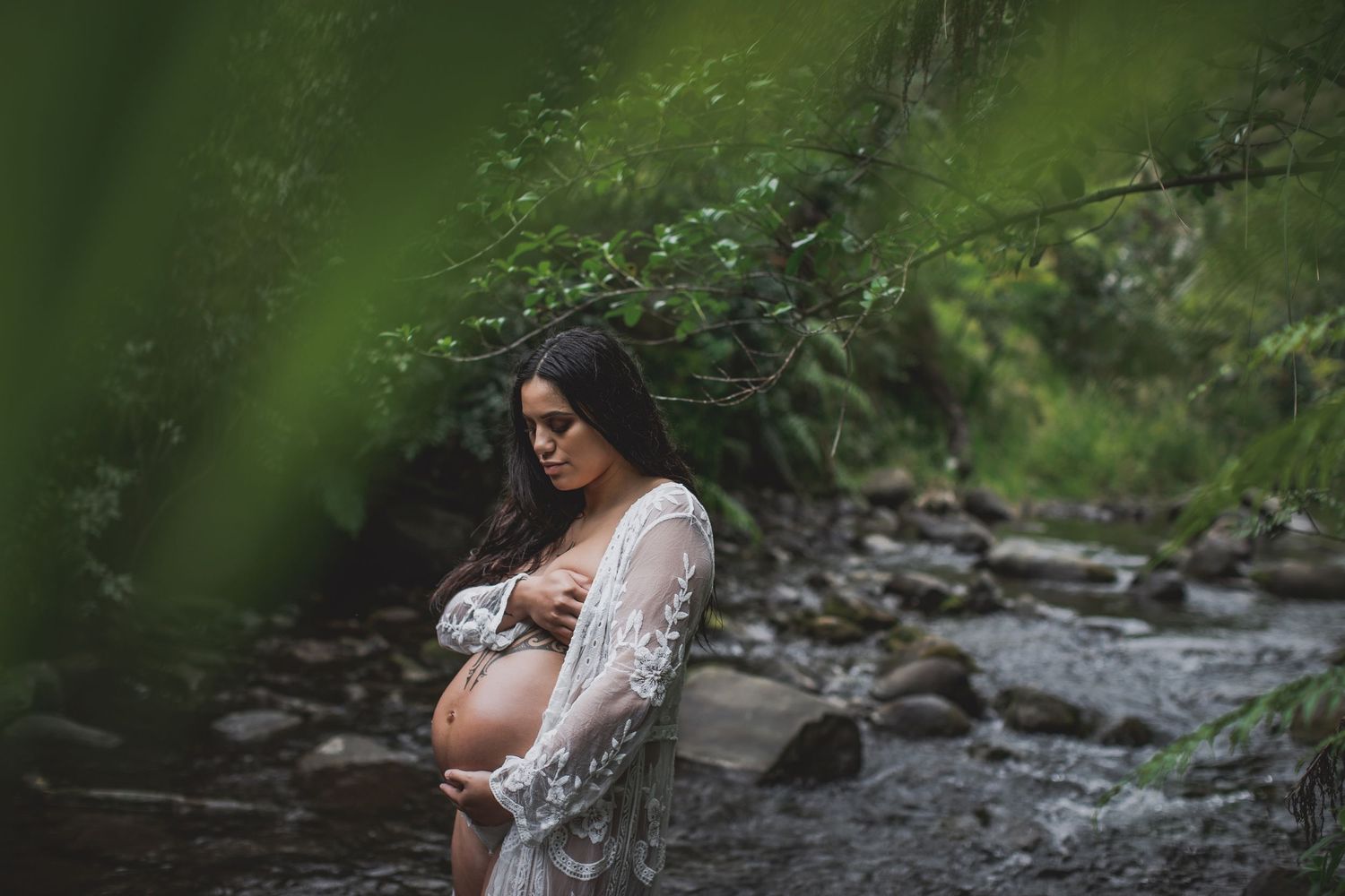 A pregnant woman in a white lace dress stands by a stream in a peaceful forest setting. Cambridge Waikato NZ Photographer. White Locket