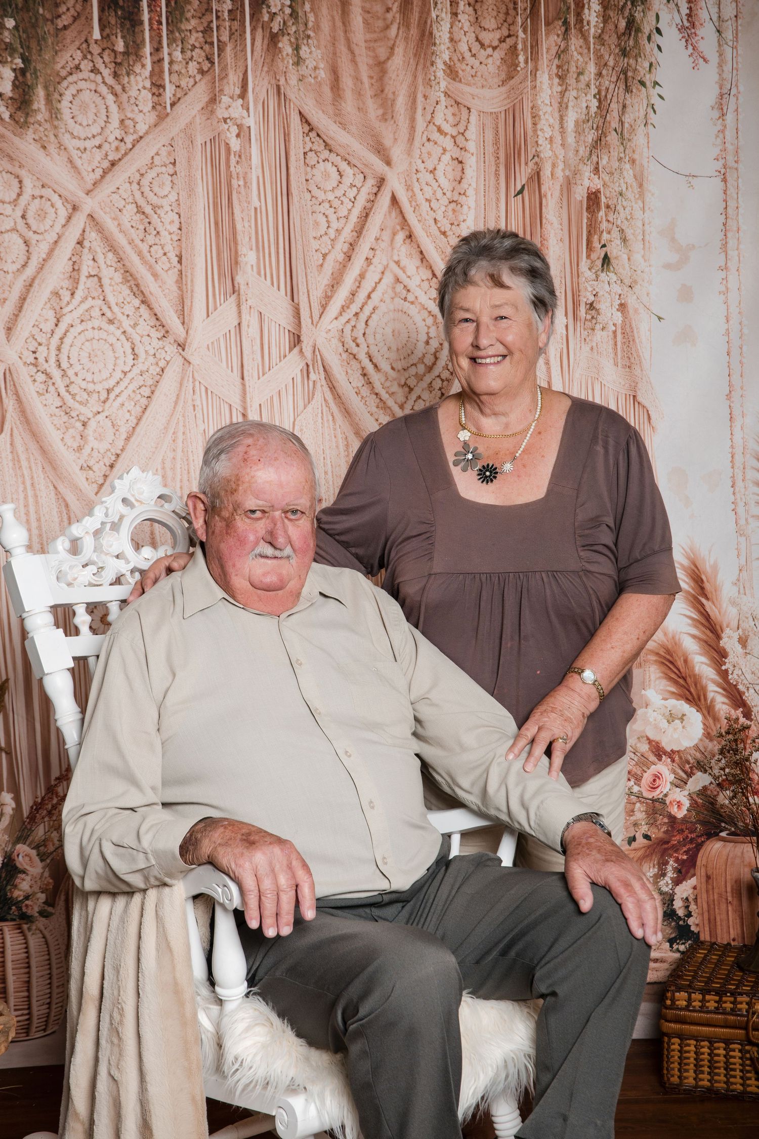 An elderly couple poses together against a macrame backdrop with decorative floral accents in a vintage style portrait.