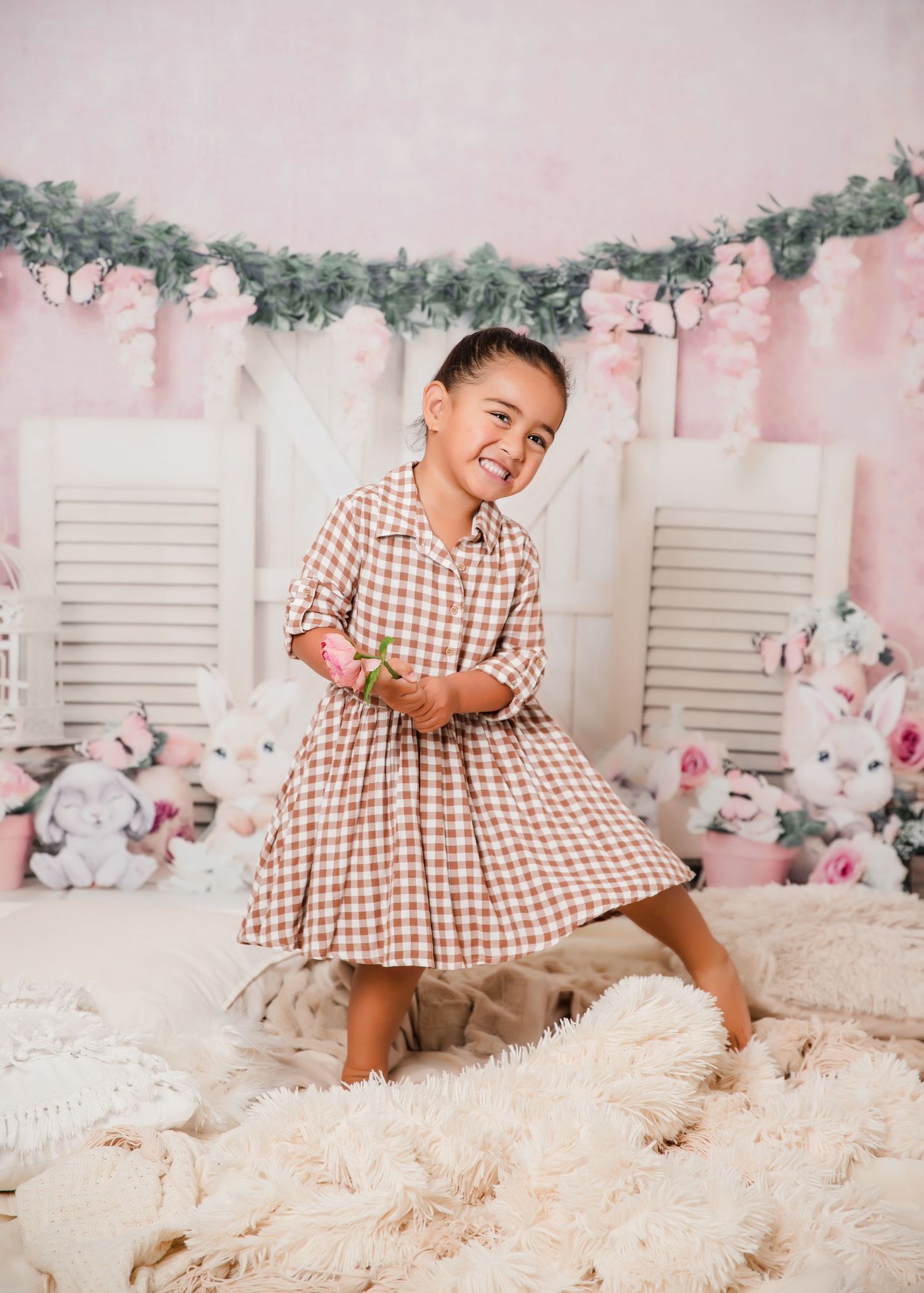 A child in a brown gingham dress twirls on a plush cream rug against a pink backdrop with floral garlands and shutters.