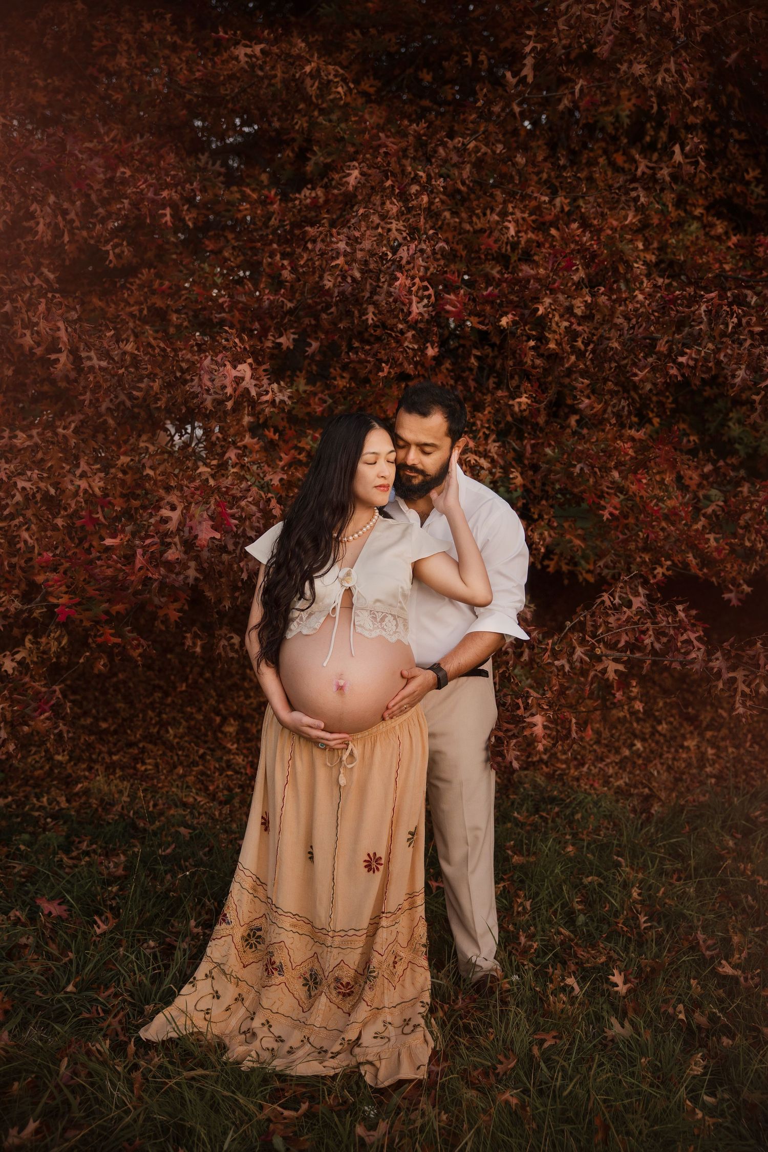 Couple sharing a tender maternity moment outdoors, surrounded by vibrant red autumn foliage.