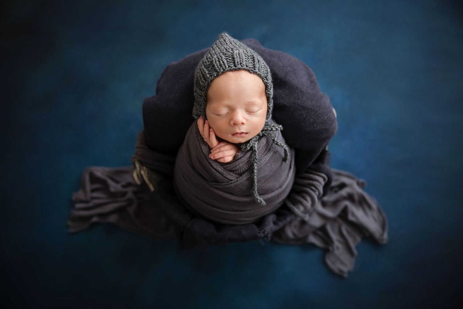 Newborn baby swaddled in dark gray wrap wearing knit bonnet, sleeping peacefully on blue textured backdrop.