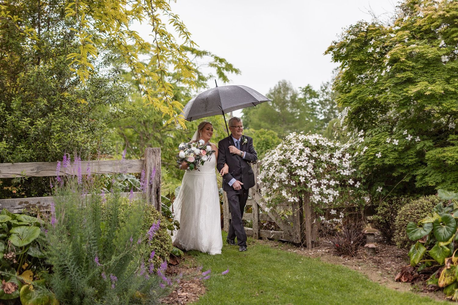 Wedding party walking along a rustic garden path during an outdoor autumn celebration with trees and umbrellas.