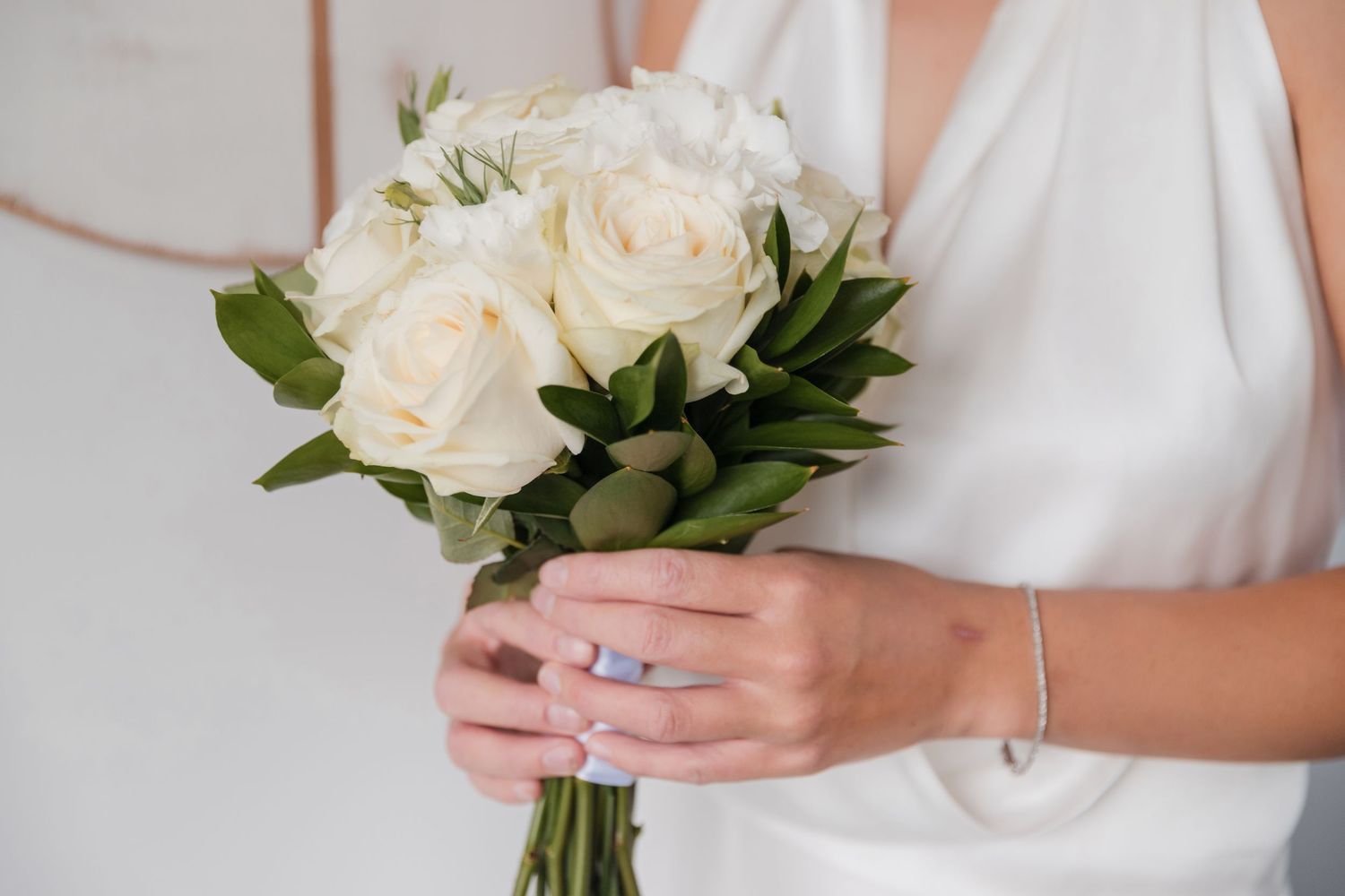 A white rose bridal bouquet is held gently against a flowing white dress in an elegant wedding photograph.