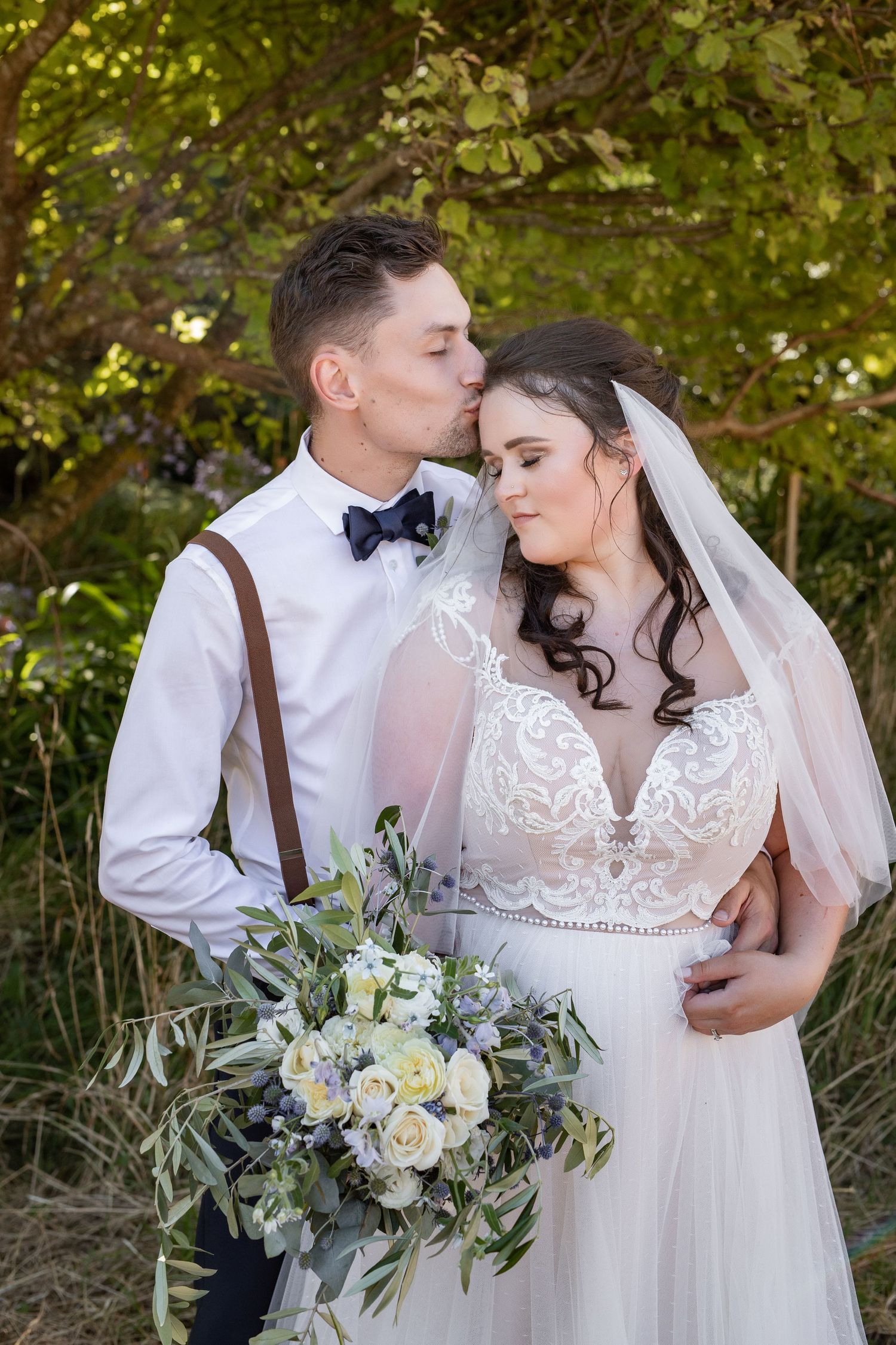 A romantic wedding couple poses outdoors with a bridal bouquet featuring white roses and greenery.
