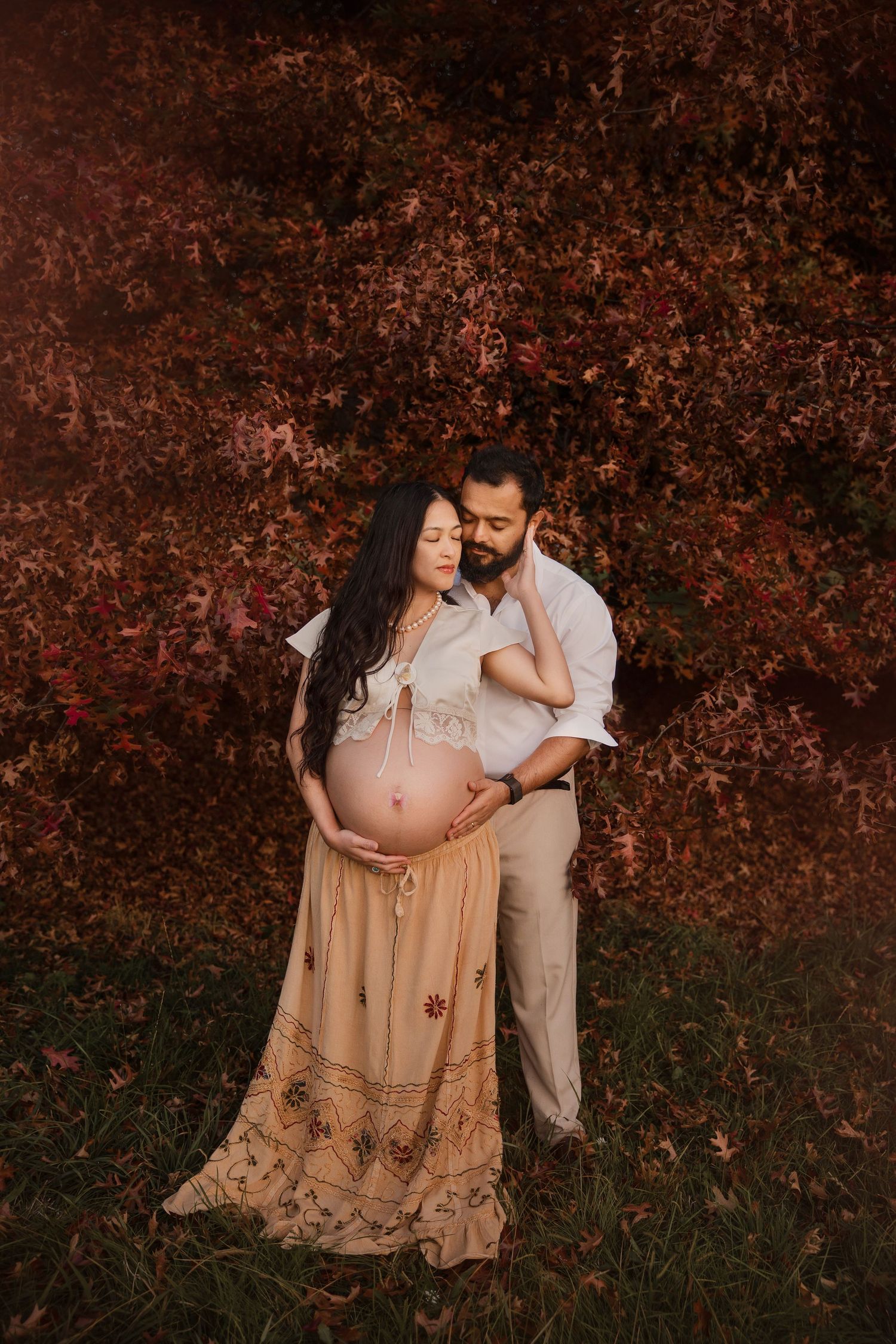 Expectant couple embracing in a maternity photoshoot, the pregnant woman in a cream boho skirt and top, surrounded by red autumn foliage.
