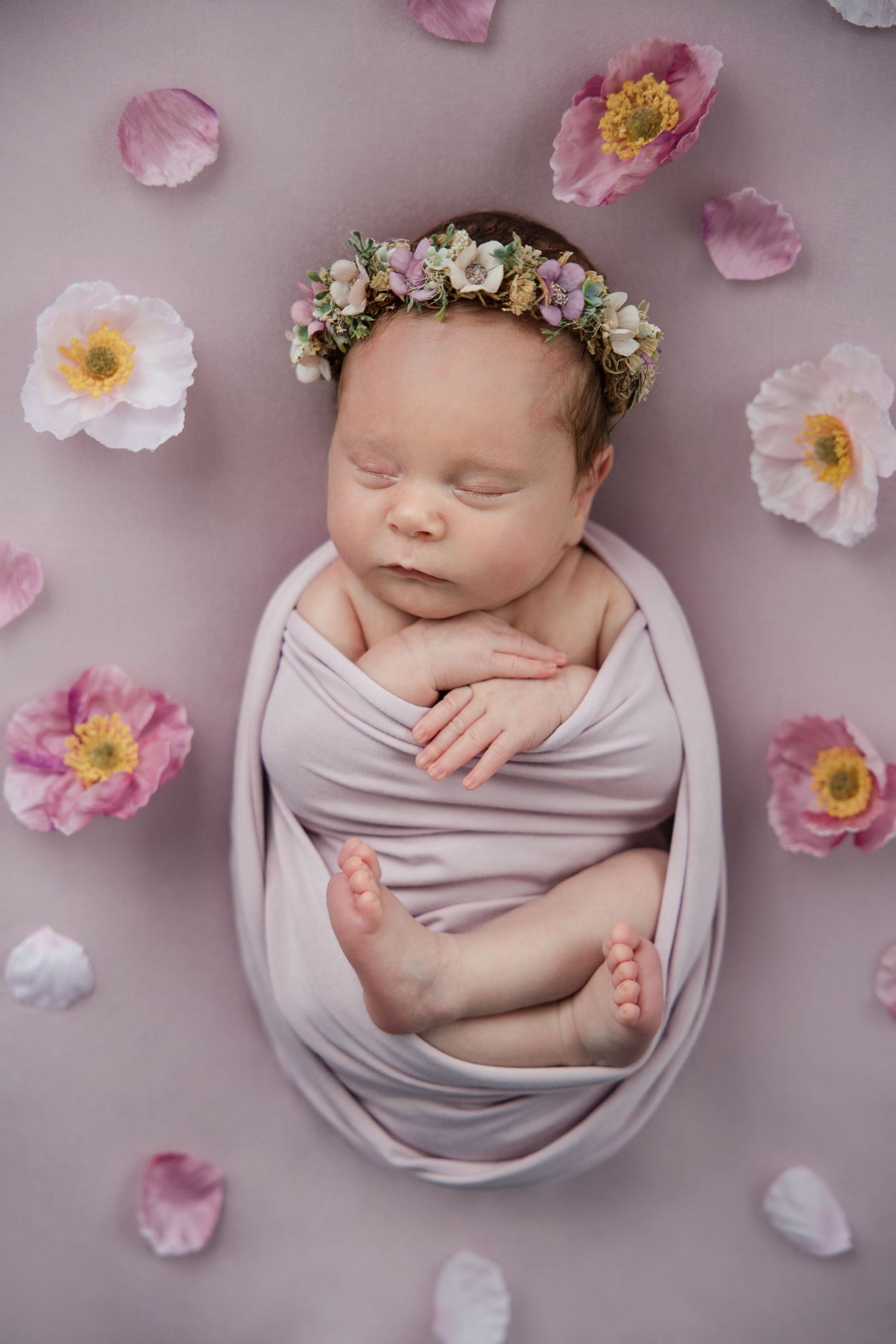 Sleeping newborn wrapped in soft gray fabric surrounded by delicate pink flower petals and wearing a floral crown.