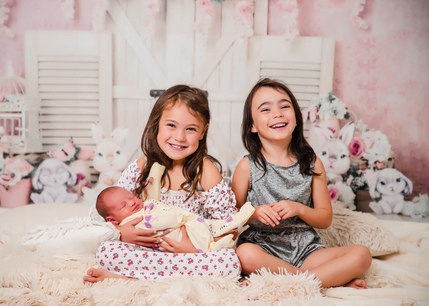 Children sitting together on a plush carpet with floral backdrop and white shutters in a pink-toned studio setting.