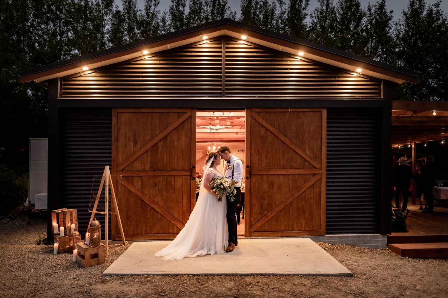 A romantic wedding couple stands in front of illuminated wooden barn doors at dusk, creating a warm glowing atmosphere.