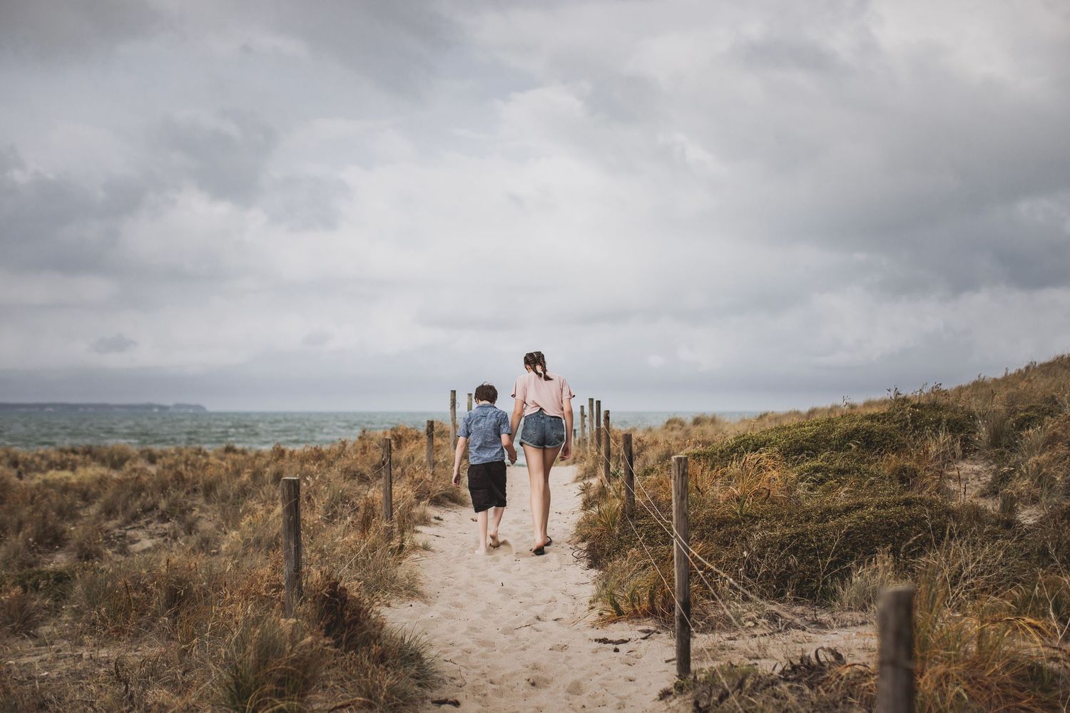 Siblings walking along sandy beach pathway through coastal dunes under cloudy sky.