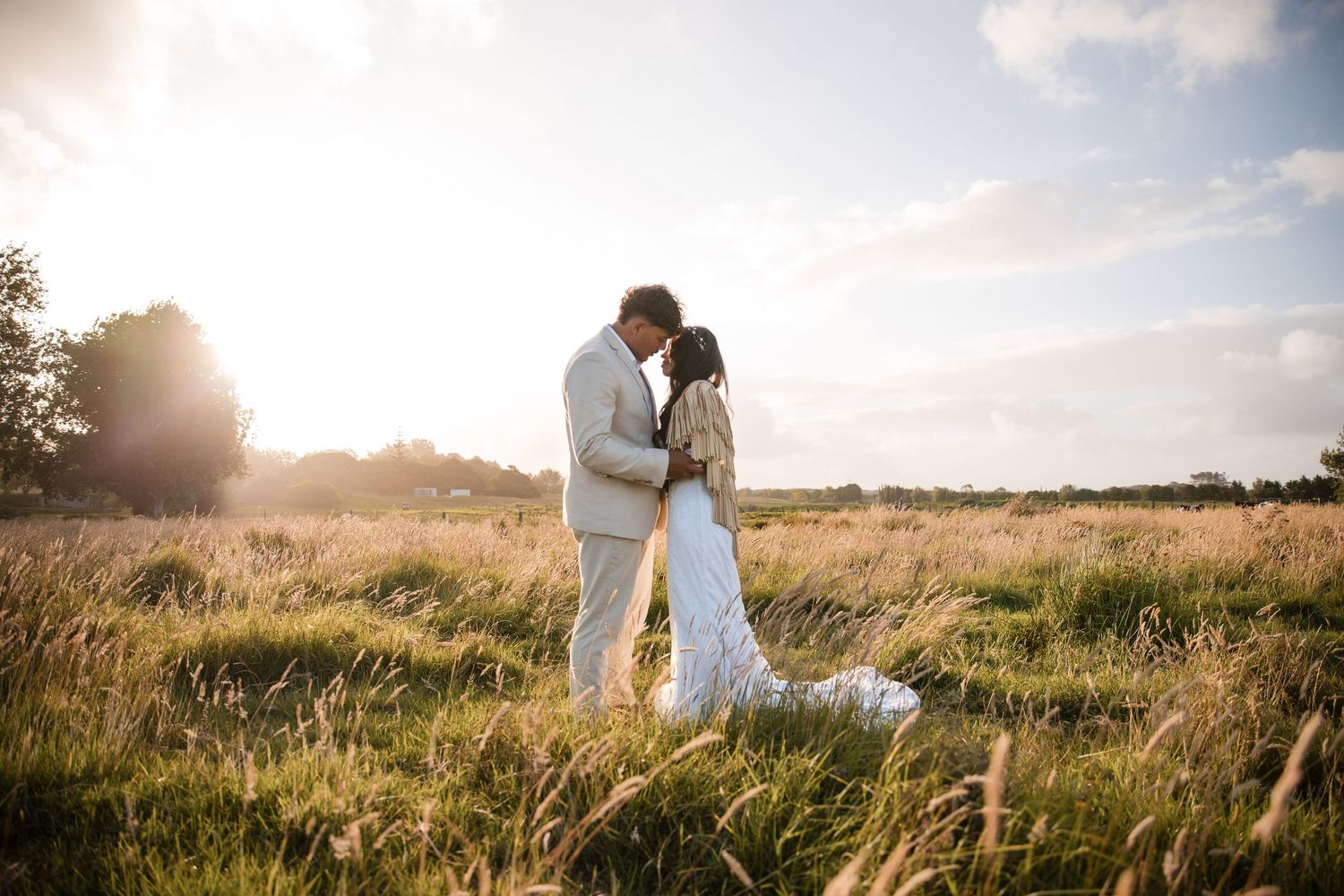Romantic sunset photo of newlyweds embracing in tall grass with golden sunlight behind them.