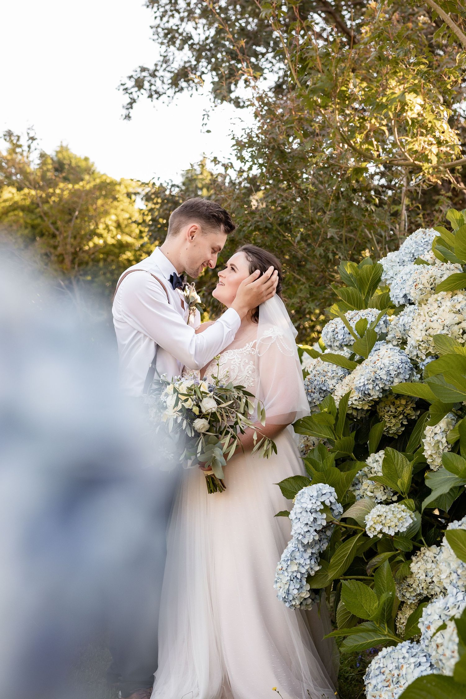 A romantic wedding couple embraces in a garden setting with blooming white hydrangeas and soft golden sunlight.