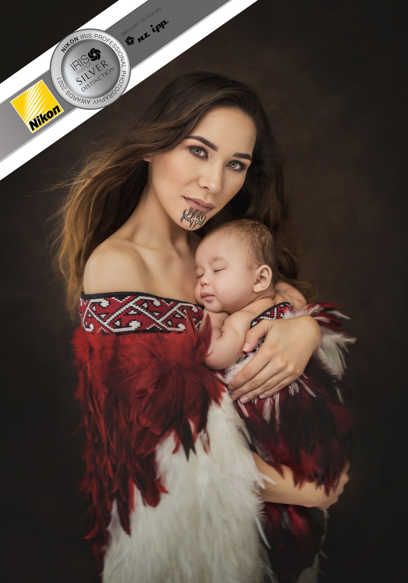 A gentle maternal portrait featuring an off-shoulder red and white dress with tribal pattern and a sleeping infant.
