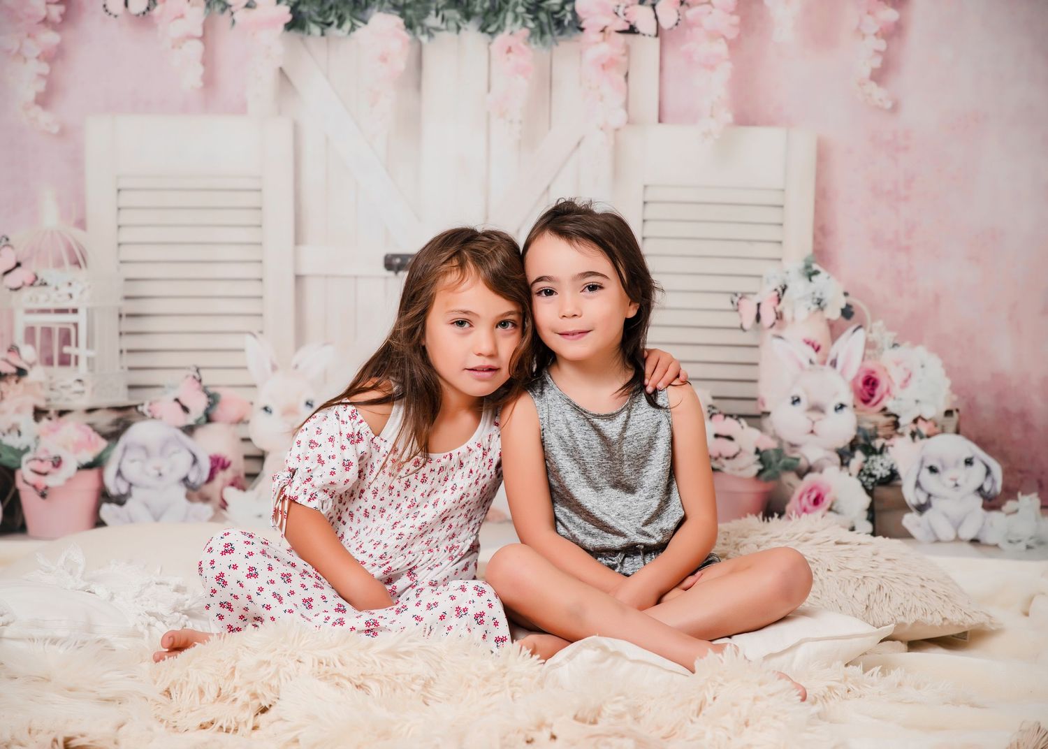 Two children sit together on a fluffy rug against a pink floral backdrop with white shutters and flowers.
